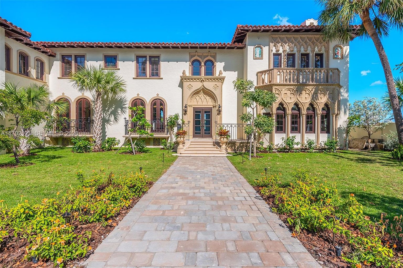 This grand Mediterranean-style estate features a symmetrical facade with intricate stone detailing, arched windows, and a prominent central entryway. A paved stone walkway leads directly to the front door, flanked by manicured lawns and lush landscaping. The property showcases a distinctive balcony with ornate stone balustrades and a classic red-tiled roof, creating an impression of luxury and architectural elegance.