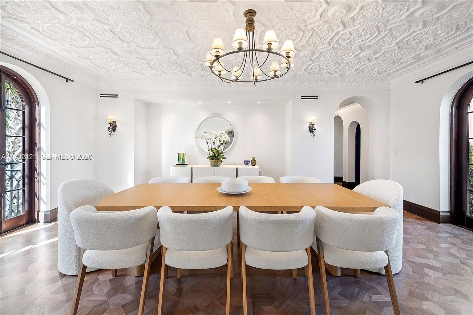 This elegant dining room features a large light wood table surrounded by eight upholstered chairs with curved backs. The space is defined by a highly detailed, ornate white ceiling, a central chandelier with multiple lights, and dark wood herringbone flooring. A sideboard sits against the back wall beneath a circular mirror, flanked by two wall-mounted sconces, creating a sophisticated and symmetrical atmosphere.