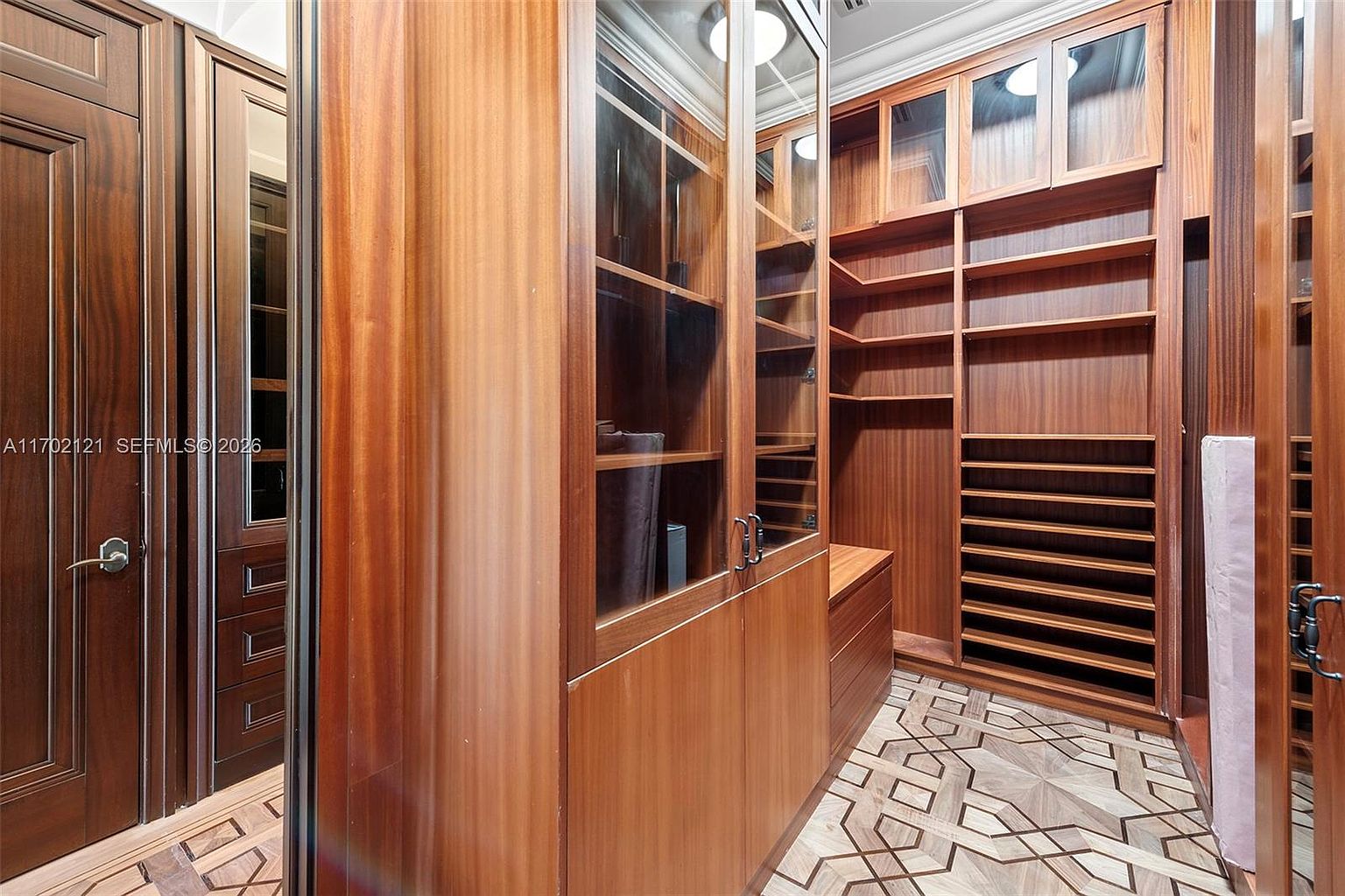 This sophisticated walk-in closet features extensive custom cabinetry crafted from rich, warm-toned wood with integrated glass-fronted doors and open shelving. The space is anchored by a unique geometric patterned tile floor, creating a luxurious and organized aesthetic. The perspective looks into the storage area, highlighting the high-end millwork and thoughtful design suitable for a high-end residential property.