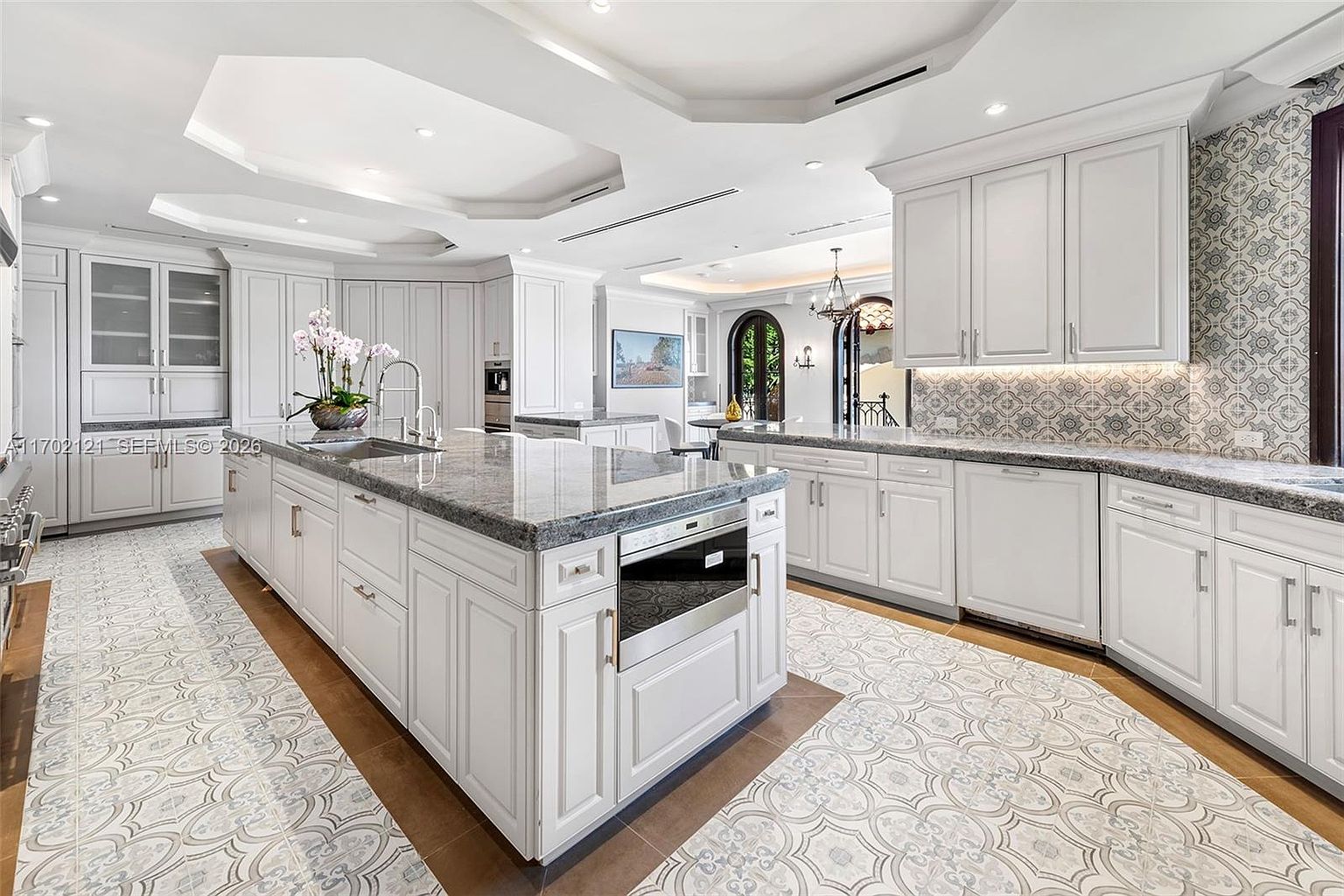 This spacious, high-end kitchen features white cabinetry, a large central island with a stone countertop, and a built-in microwave drawer. The floor is accented with decorative patterned tile inlays set into a neutral tile base, while a matching patterned tile backsplash adds visual interest behind the counters. The perspective is a wide-angle shot from the kitchen floor, showcasing the expansive layout, recessed lighting, and a glimpse into the adjacent dining area.
