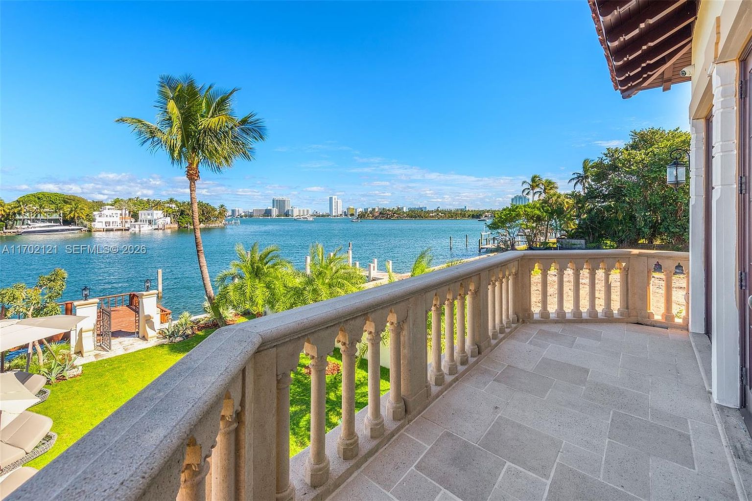 This elevated stone balcony offers a sweeping, panoramic view of a tranquil bay and the distant city skyline. The space features a classic stone balustrade and tiled flooring, overlooking a lush green lawn, a private dock, and a single palm tree. The perspective is from the balcony looking outward, capturing a serene and luxurious waterfront atmosphere.