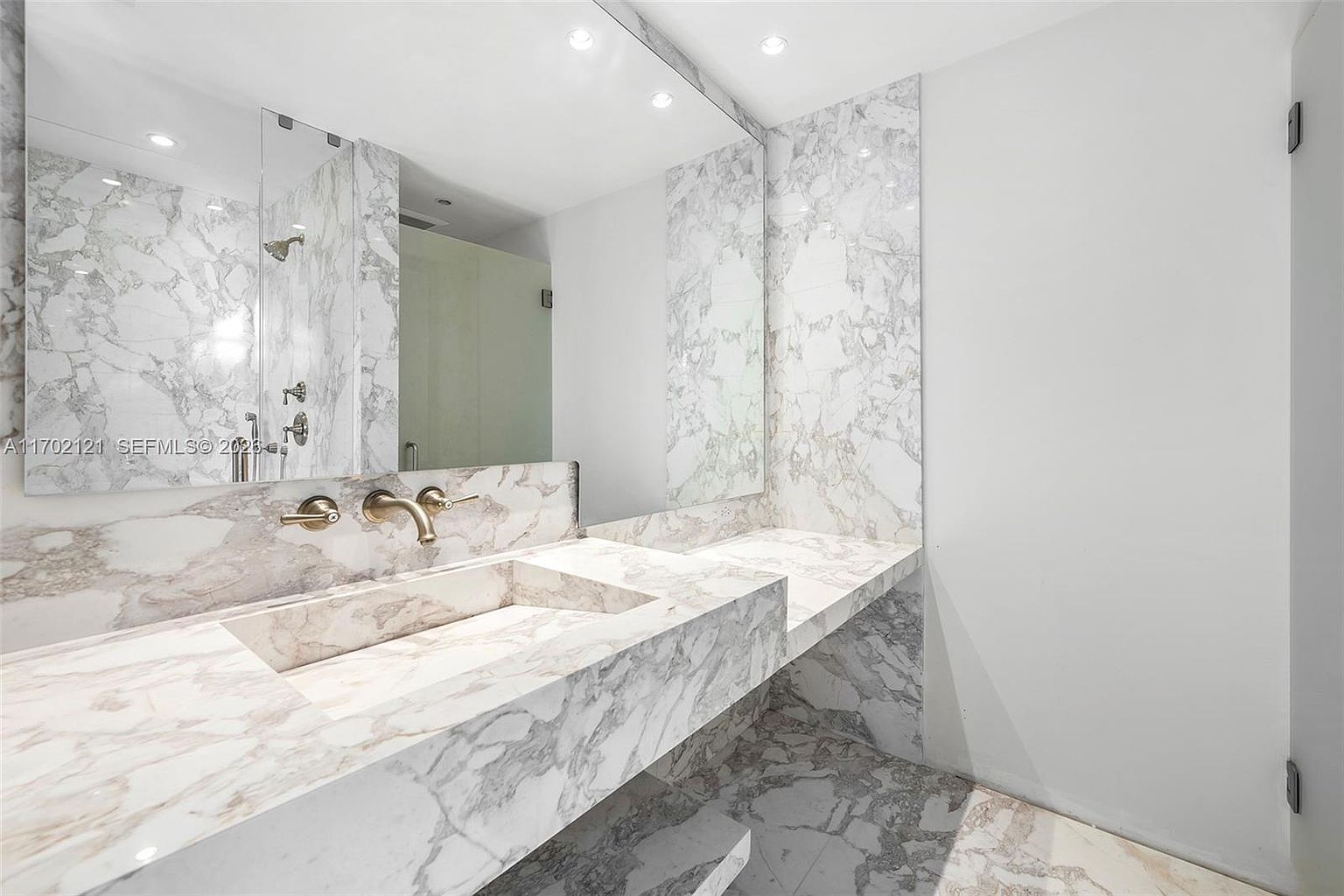 This luxurious bathroom features a striking, seamless stone vanity with an integrated sink and gold-toned fixtures. A large, frameless mirror spans the wall above the vanity, reflecting the adjacent walk-in shower area. The space is finished with matching stone surfaces on the walls and floor, creating a cohesive and high-end aesthetic.