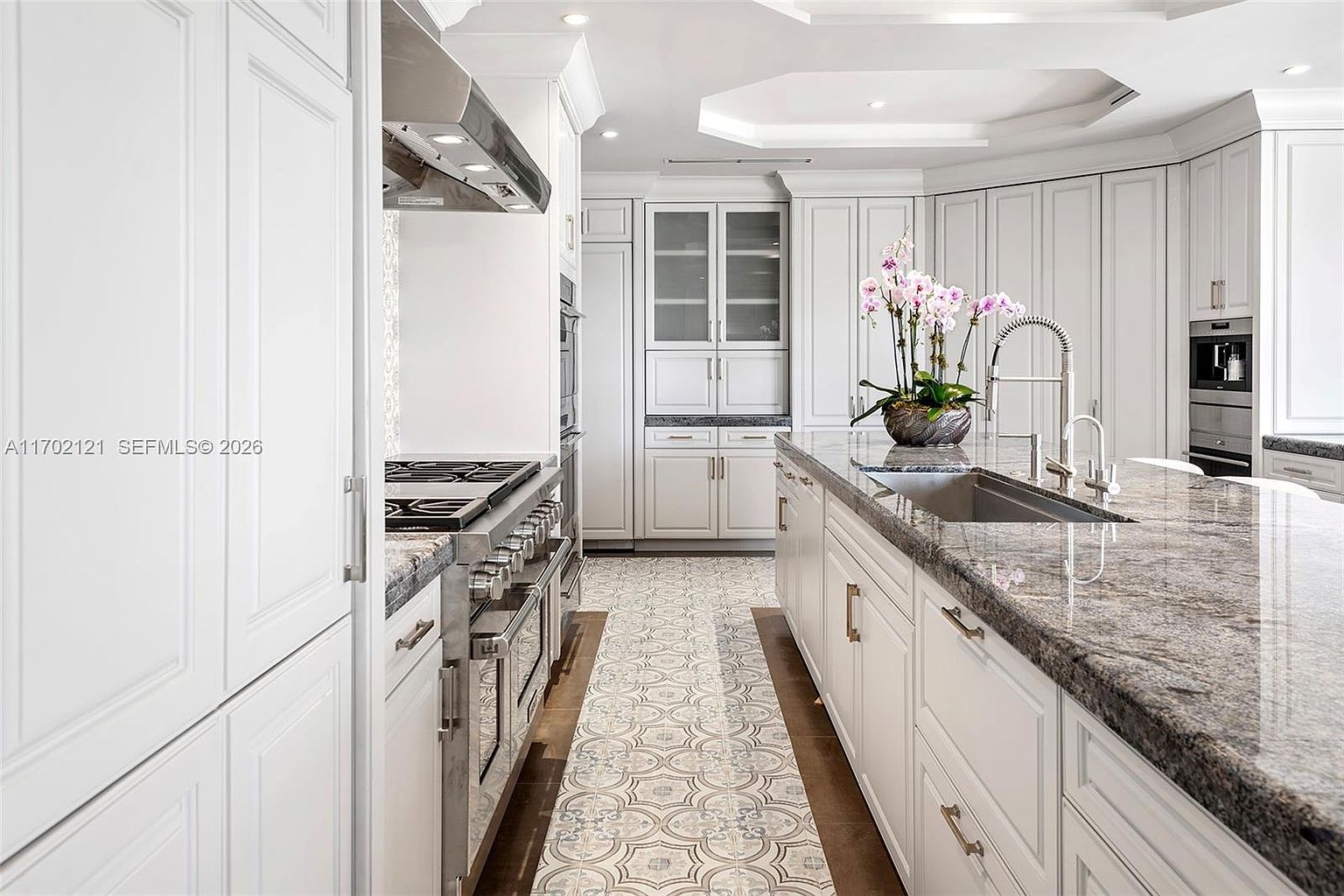 This elegant, high-end kitchen features crisp white cabinetry paired with a large central island topped with a polished stone countertop. A decorative patterned tile runner runs along the floor between the island and the professional-grade stainless steel range, creating a sophisticated focal point. The space is well-lit with recessed lighting and includes integrated appliances, offering a clean, luxurious, and functional culinary environment.
