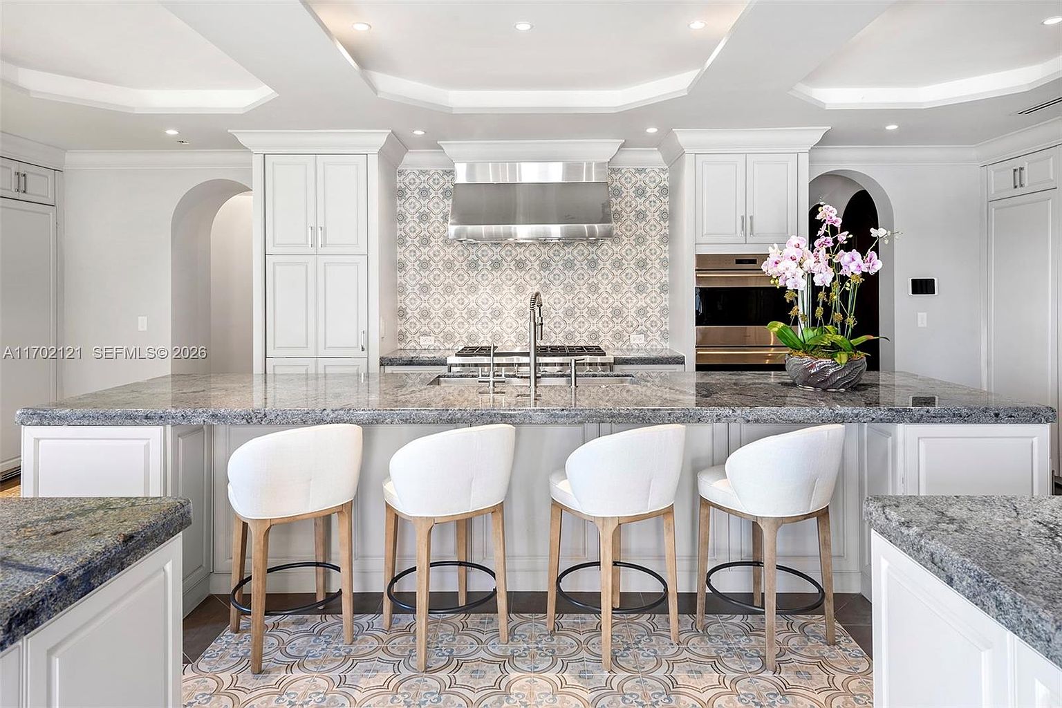 This elegant kitchen features a large central island with a dark stone countertop and four light-toned stools with white upholstered seats. The space is anchored by white cabinetry, a stainless steel range hood, and a decorative patterned tile backsplash that coordinates with the flooring. The perspective is a straight-on, eye-level shot that highlights the symmetrical layout and sophisticated design of the culinary space.