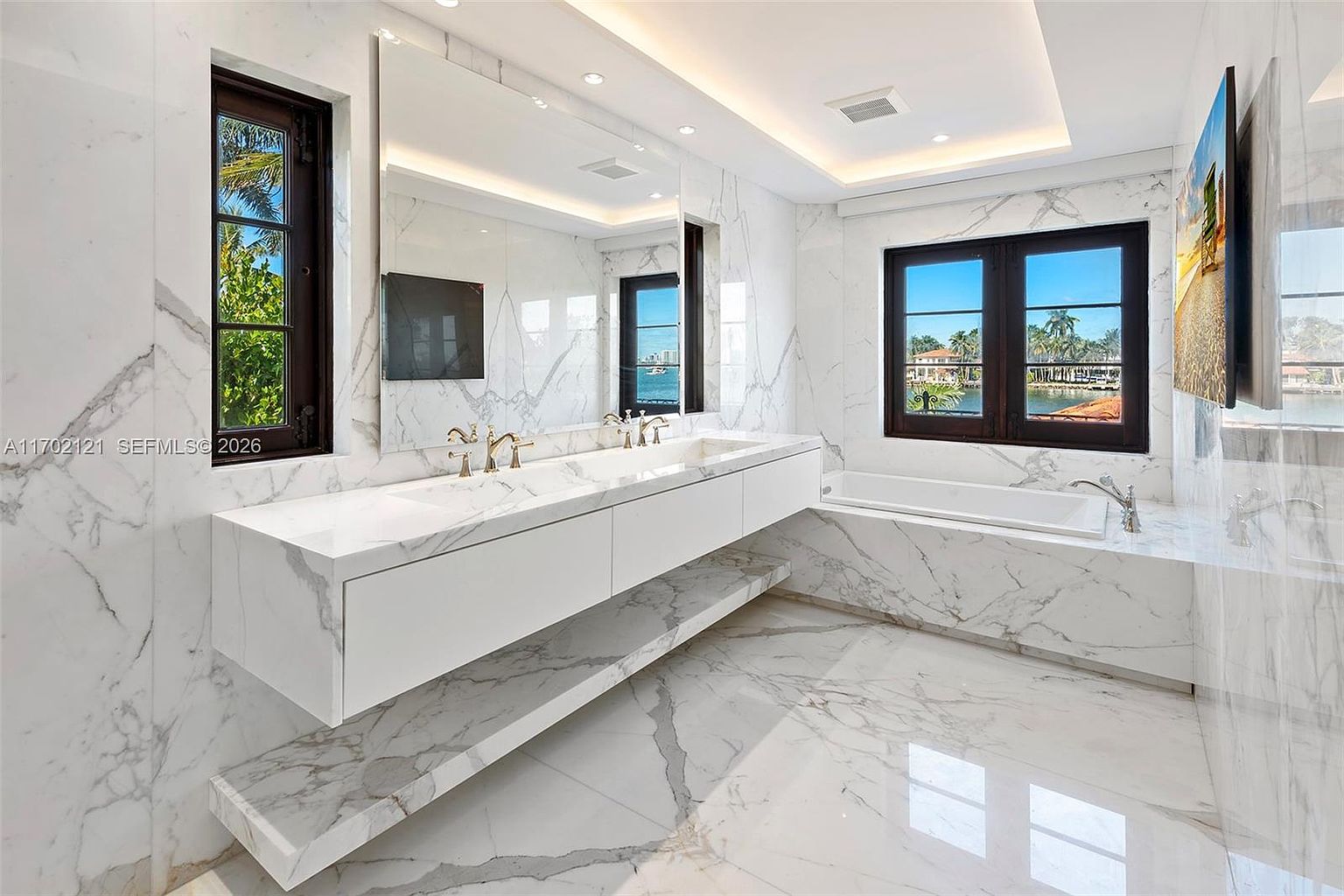 This luxurious primary bathroom features extensive use of white stone with dramatic grey veining on the walls and floors. The space includes a long, floating double vanity with integrated sinks, a large mirror, and a deep soaking tub positioned beneath a window with a water view. The room is illuminated by recessed lighting and natural light, creating a bright, high-end, and serene atmosphere.