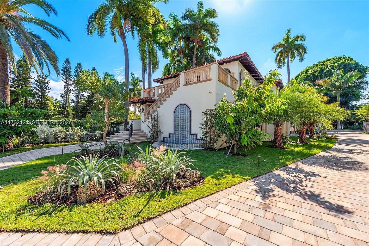 This side view of a Mediterranean-style home showcases a distinctive exterior staircase leading to an upper-level balcony with a balustrade. A decorative arched wall feature with blue patterned tiles serves as a focal point on the lower level, set against a lush landscape of palm trees and manicured lawn. The scene is captured from a low angle on a paved driveway, emphasizing the architectural character and tropical surroundings of the property.