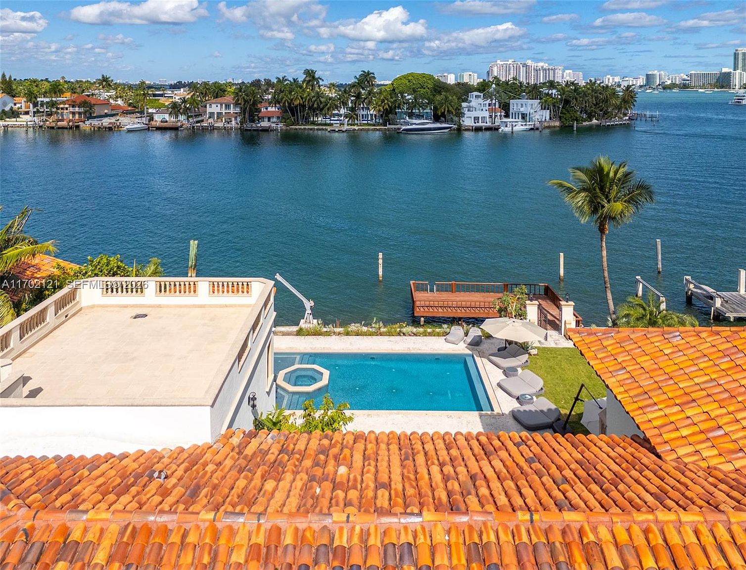 This high-angle aerial view captures a luxurious waterfront property featuring a rectangular swimming pool set on a stone patio. The foreground showcases a classic terracotta-tiled roof, while the backyard leads directly to a private wooden dock extending into a calm, expansive bay. In the distance, a scenic shoreline with lush palm trees and multi-story residential buildings completes the serene, upscale coastal atmosphere.