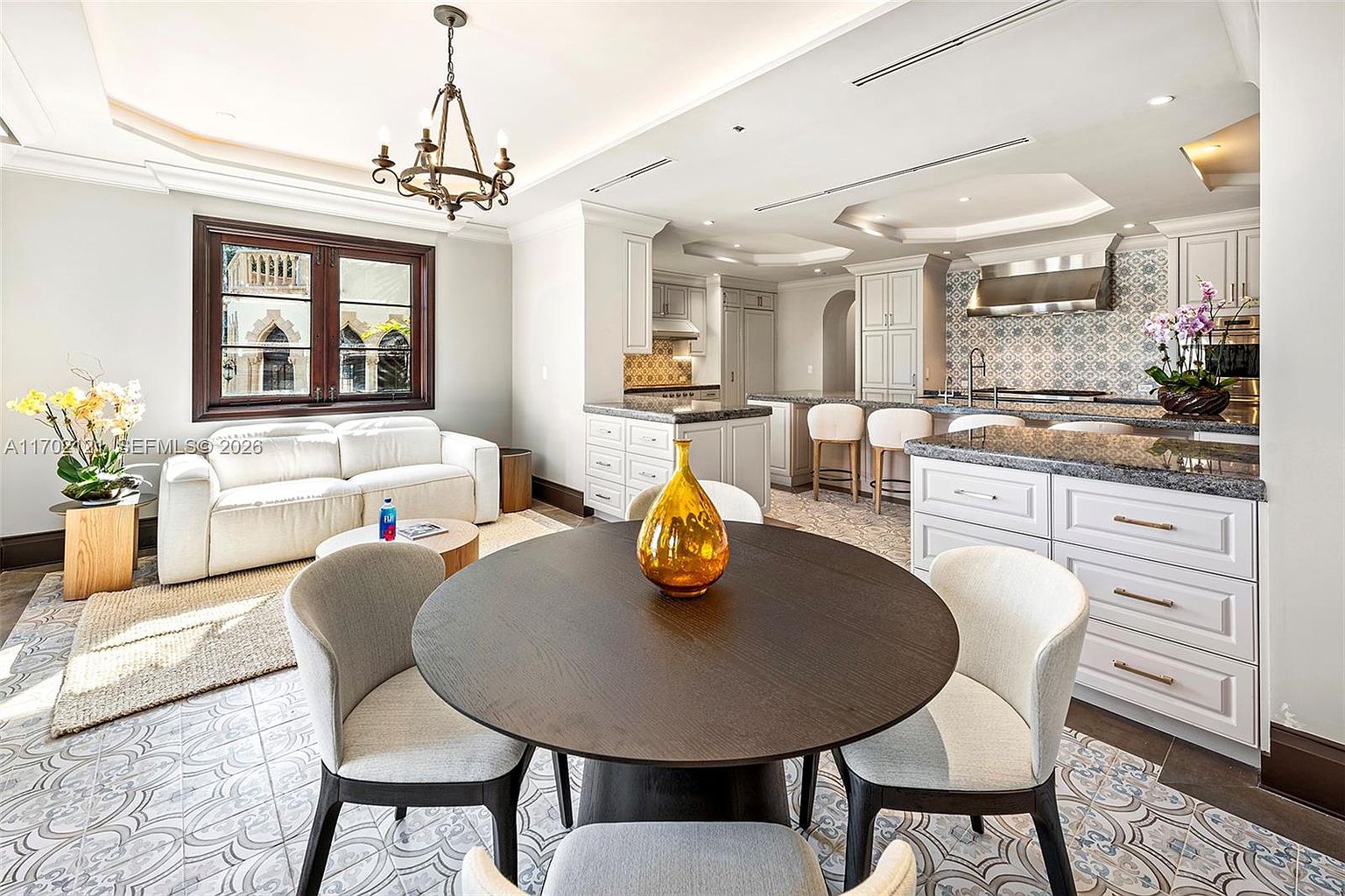 This open-concept space seamlessly blends a dining area with a spacious, high-end kitchen featuring white cabinetry and patterned tile backsplashes. A dark circular dining table sits in the foreground, while the kitchen island provides additional seating and a central hub for the room. The area is illuminated by a decorative chandelier and recessed lighting, creating a bright and sophisticated atmosphere.