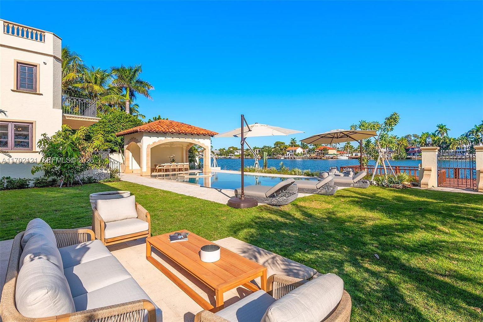 This expansive waterfront backyard features a well-manicured lawn, a private swimming pool, and a covered poolside cabana. In the foreground, a comfortable outdoor seating area with a wooden coffee table and plush sofas provides a perfect vantage point for enjoying the serene water views. The scene is bathed in bright, natural sunlight under a clear blue sky, highlighting the resort-like atmosphere of the property.