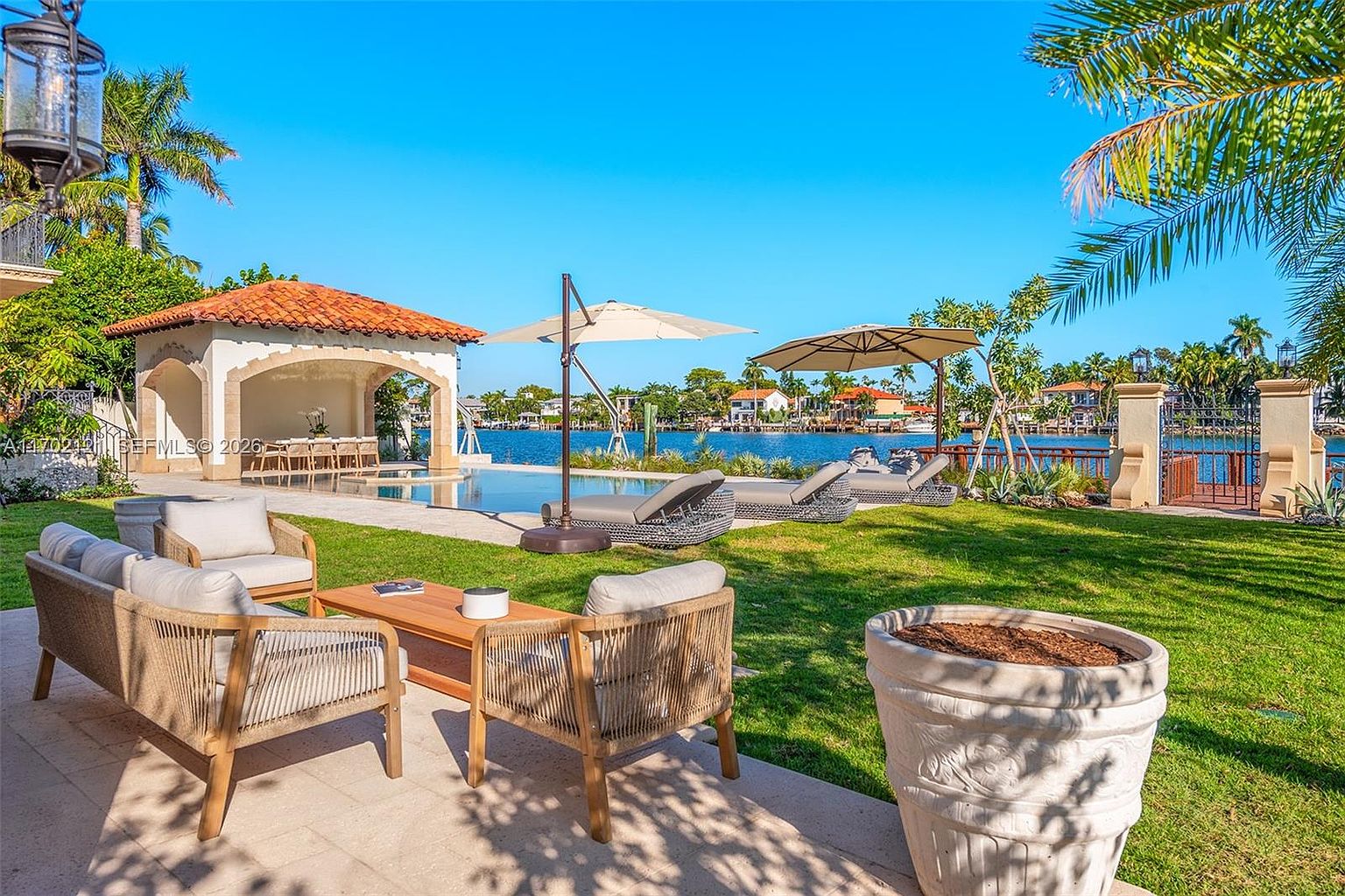 This expansive waterfront backyard features a pristine infinity-edge pool and a covered poolside pavilion with a terracotta-tiled roof. The manicured lawn is furnished with comfortable outdoor seating, including lounge chairs and a conversational set, all overlooking a serene canal. The scene is captured from a ground-level perspective, emphasizing the luxurious outdoor living space and the tranquil water views.
