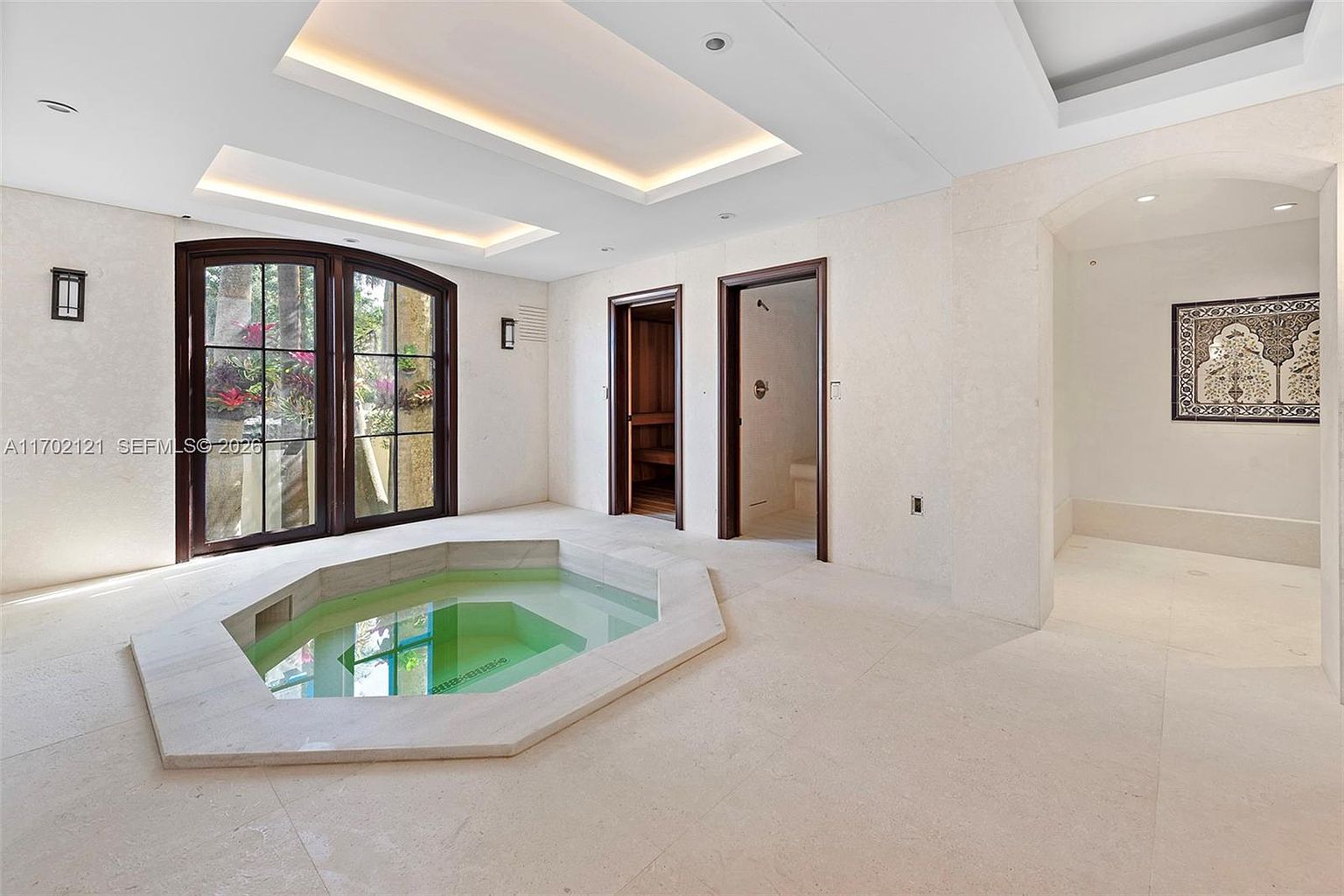 This luxurious spa-inspired room features a large, octagonal sunken tub as the central focal point, set into a light-toned stone surround. The space includes a sauna and a separate shower stall, both accessed through dark wood-framed doorways, and is illuminated by recessed lighting within a tiered ceiling design. A decorative, intricate tile mural adorns the wall on the right, adding a touch of elegance to the minimalist, light-colored stone aesthetic.