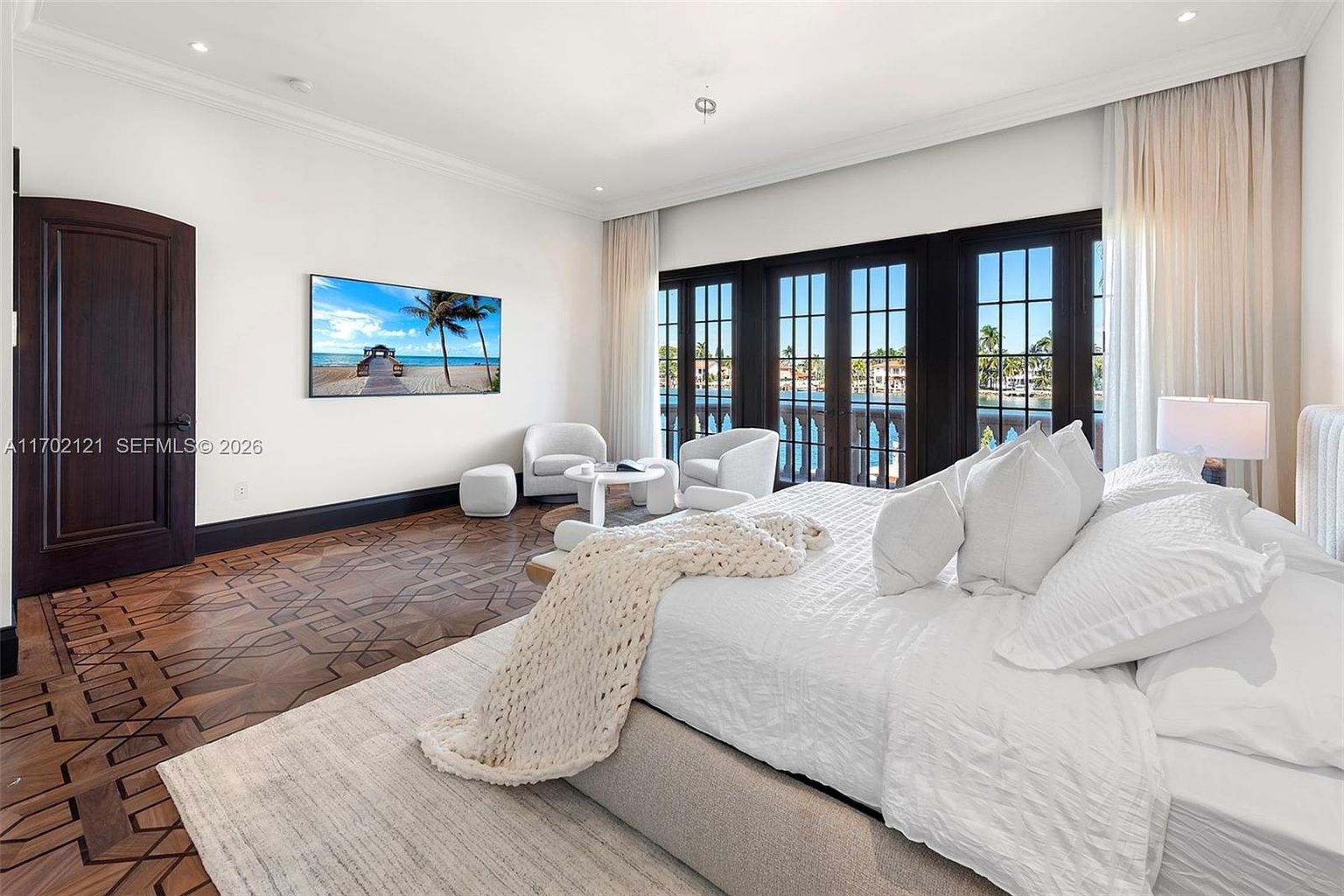 This spacious primary bedroom features intricate dark wood parquet flooring and a large, plush bed dressed in white linens. A seating area with two armchairs and a small table is positioned near a wall of tall, multi-paned glass doors that open to a balcony with water views. The room is bright and airy, with neutral walls and a wall-mounted television, creating a serene and luxurious atmosphere.
