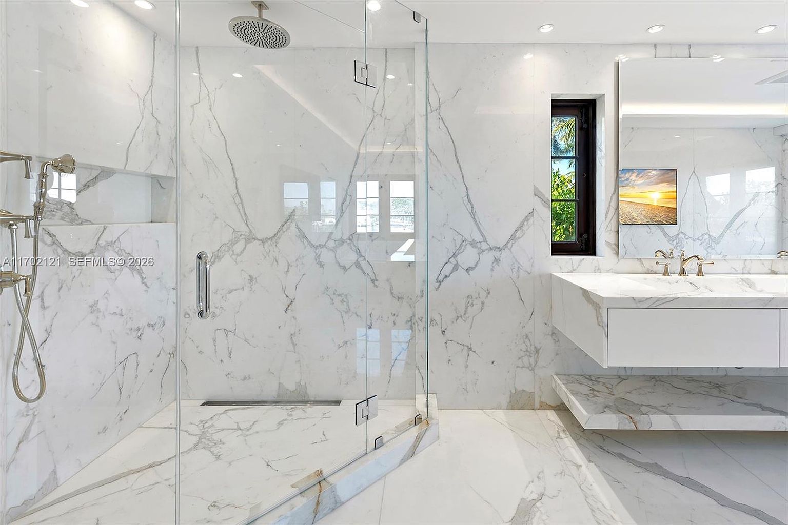 This luxurious primary bathroom features extensive use of white stone with dramatic grey veining throughout the walls, shower enclosure, and vanity surfaces. The space includes a glass-enclosed walk-in shower with a rainfall showerhead and a floating vanity with dual faucets, all complemented by a large mirror and a narrow window that brings in natural light. The overall aesthetic is sleek, modern, and high-end, emphasizing clean lines and a bright, airy atmosphere.