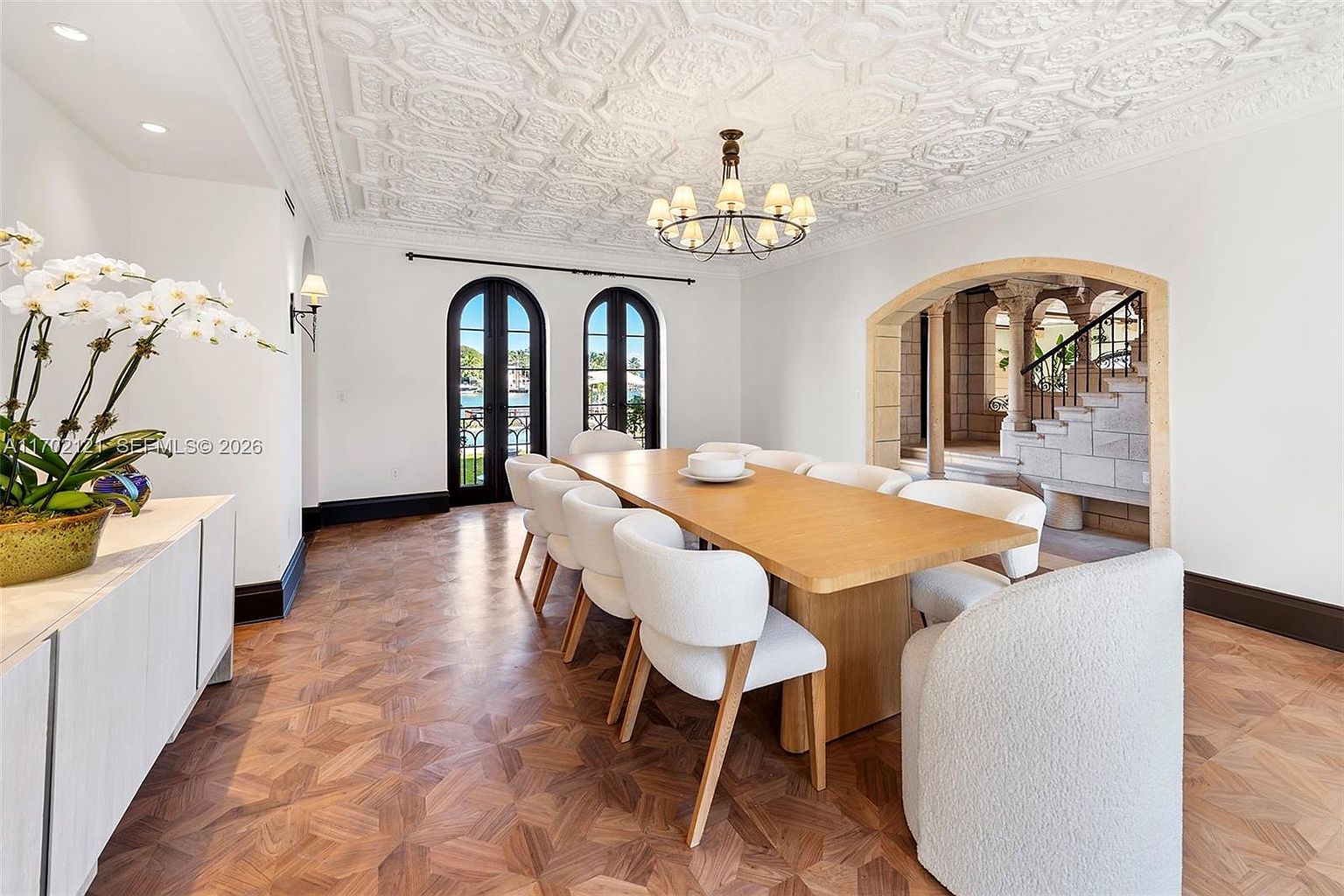This elegant dining room features a long, light wood table surrounded by white upholstered chairs, set beneath a classic chandelier and an intricate, patterned ceiling. The room showcases rich, patterned wood flooring and arched glass doors that provide a view of the outdoors. An arched stone opening leads to a staircase, adding a sophisticated architectural element to the space.