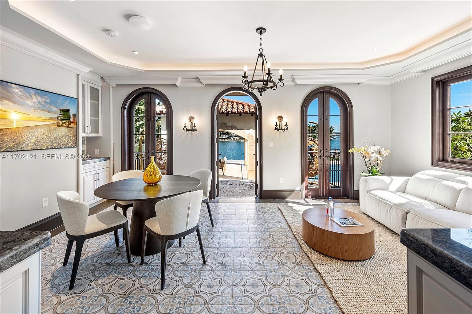 This elegant open-concept space seamlessly blends a dining area with a cozy living nook, featuring intricate patterned tile flooring throughout. The room is anchored by a dark wood oval dining table and a central chandelier, while three arched glass doors provide a direct view and access to a waterfront patio. A white sofa and a low-profile coffee table define the living area, creating a sophisticated and airy atmosphere perfect for entertaining.