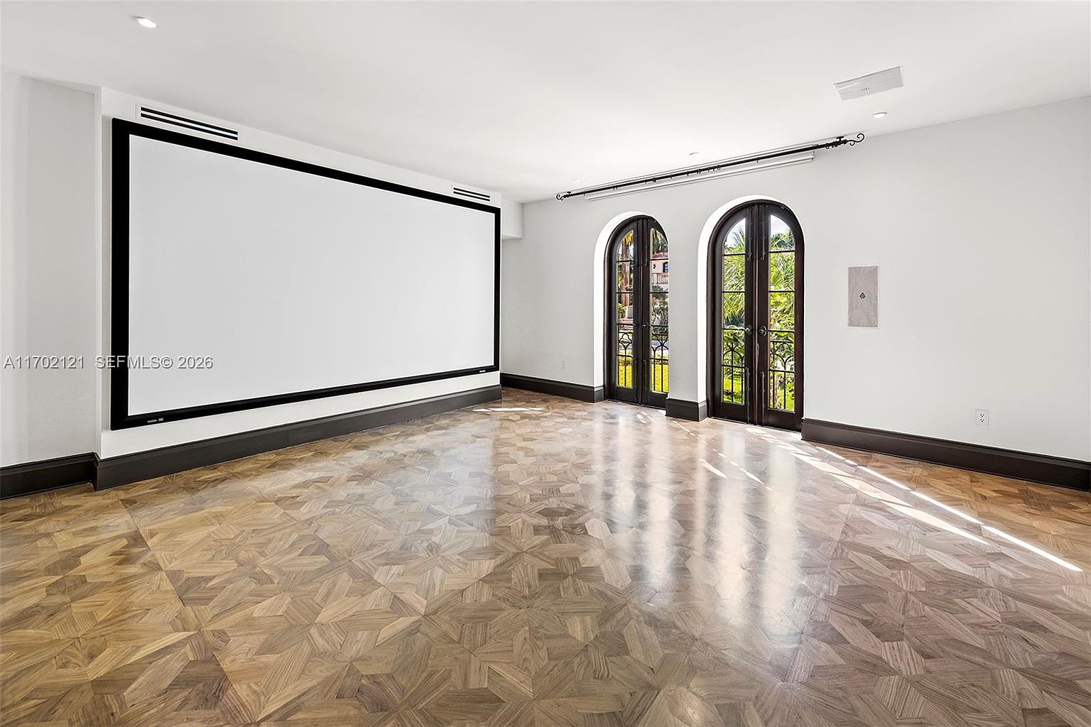 This spacious media room features a large, wall-mounted projection screen and intricate, light-toned wood parquet flooring arranged in a geometric pattern. Two arched glass doors with dark frames provide natural light and a view of the outdoors, while the room is finished with clean white walls and dark baseboards. The perspective is wide and centered, emphasizing the room's potential as a dedicated entertainment space.
