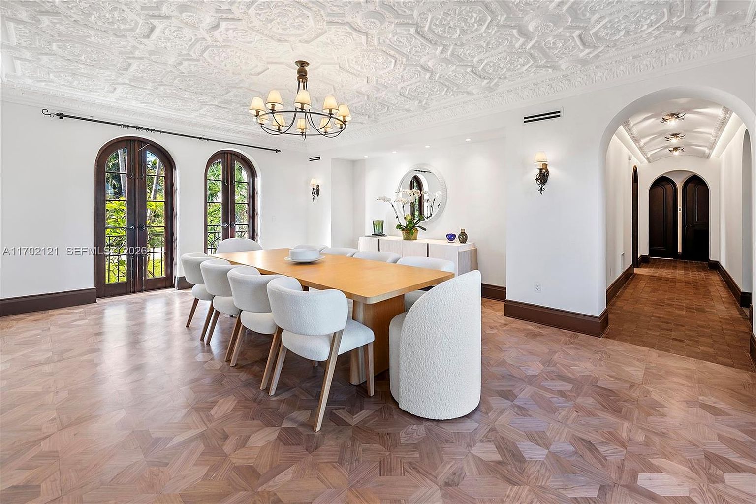 This elegant dining room features a large wooden table surrounded by eight upholstered chairs, set against a backdrop of intricate, ornate ceiling molding. The space is illuminated by a classic chandelier and wall sconces, with tall arched glass doors providing natural light and a view of the outdoors. A hallway with a barrel-vaulted ceiling leads away from the room, showcasing a sophisticated, traditional design aesthetic.
