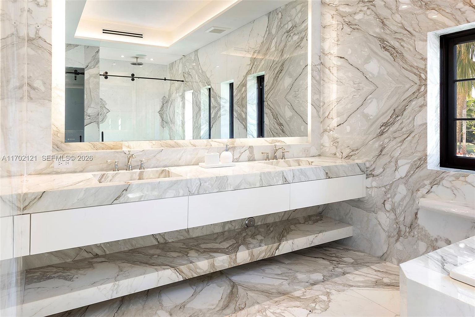 This luxurious primary bathroom features a double vanity with a long, patterned stone countertop and integrated sinks, set against a backdrop of matching stone wall cladding. A large, frameless mirror spans the width of the vanity, illuminated by integrated lighting, while a floating white cabinet provides sleek storage. The space is finished with a matching stone shelf below the vanity and a window that allows natural light to brighten the room.