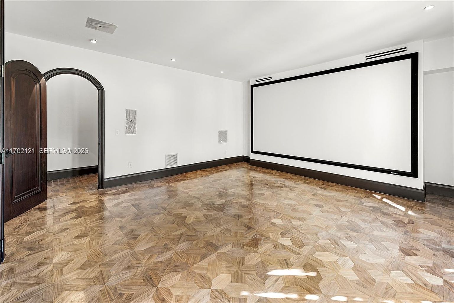 This image features a dedicated home theater room characterized by a large, wall-mounted projection screen and a distinctive geometric patterned wood floor. The room is framed by a dark-trimmed arched doorway and features recessed lighting, creating a clean and minimalist aesthetic. The perspective is from the center of the room, looking toward the screen, emphasizing the spacious and specialized nature of the space.