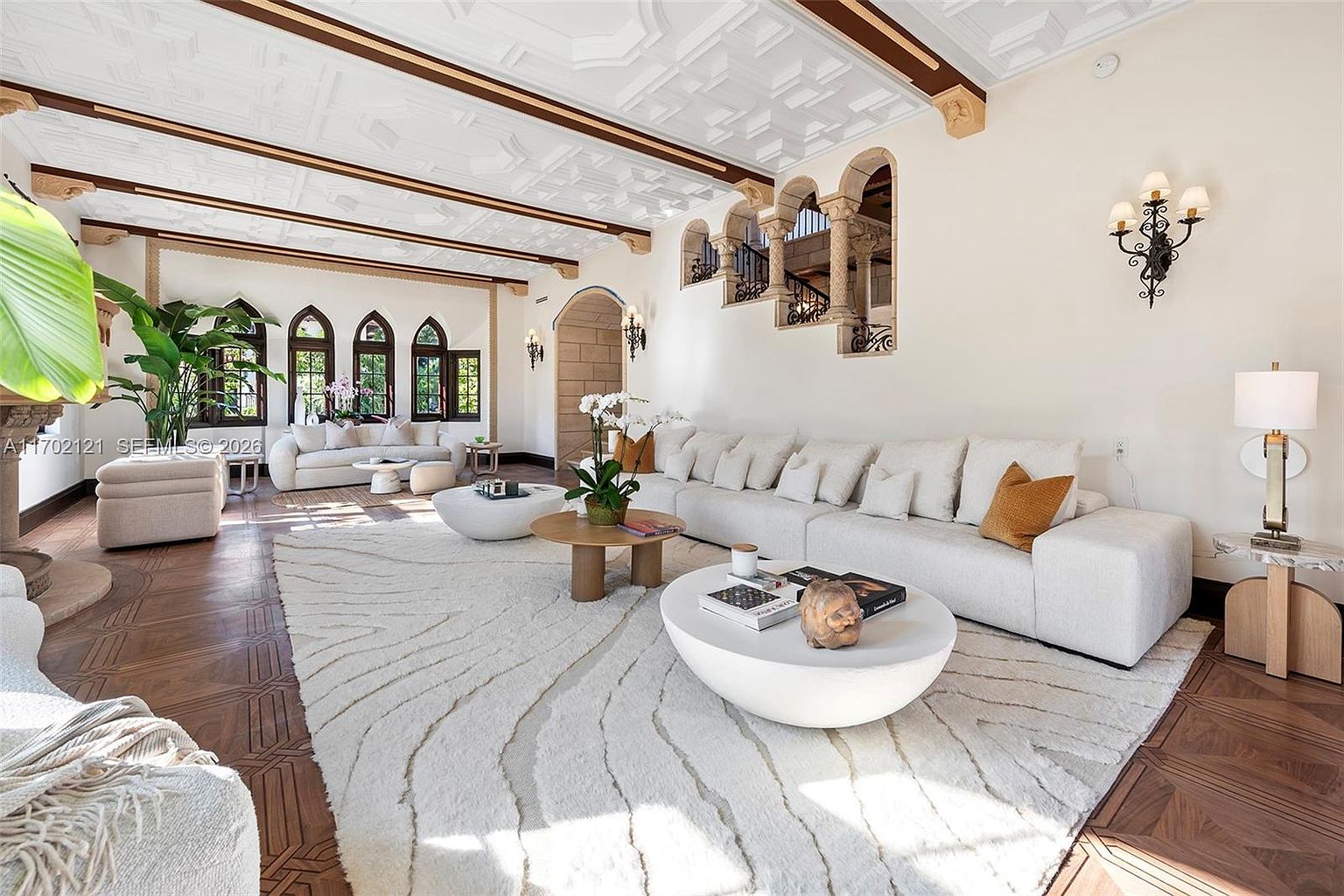 This expansive living room features a sophisticated blend of Mediterranean-inspired architectural details, including a coffered ceiling with dark wood beams and arched wall openings. The space is anchored by a large, plush white sectional sofa and a textured area rug, creating a bright and airy atmosphere. The room is accented by warm wood flooring, a sculptural coffee table, and elegant wall sconces, offering a luxurious and inviting aesthetic.
