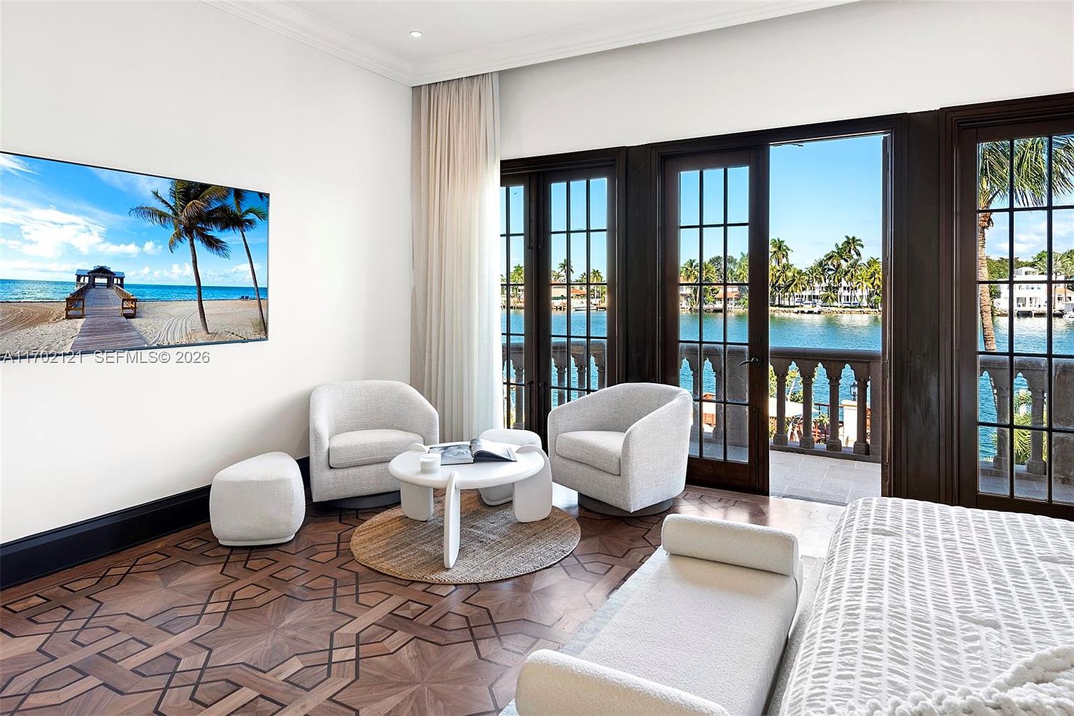 This elegant bedroom suite features intricate geometric patterned wood flooring and a cozy seating area with two light-colored armchairs and a round coffee table. Large glass doors open onto a balcony with a stone balustrade, providing a stunning, unobstructed view of the waterfront and palm trees. The room is bright and airy, with a large wall-mounted television displaying a tropical beach scene, enhancing the serene, resort-like atmosphere.