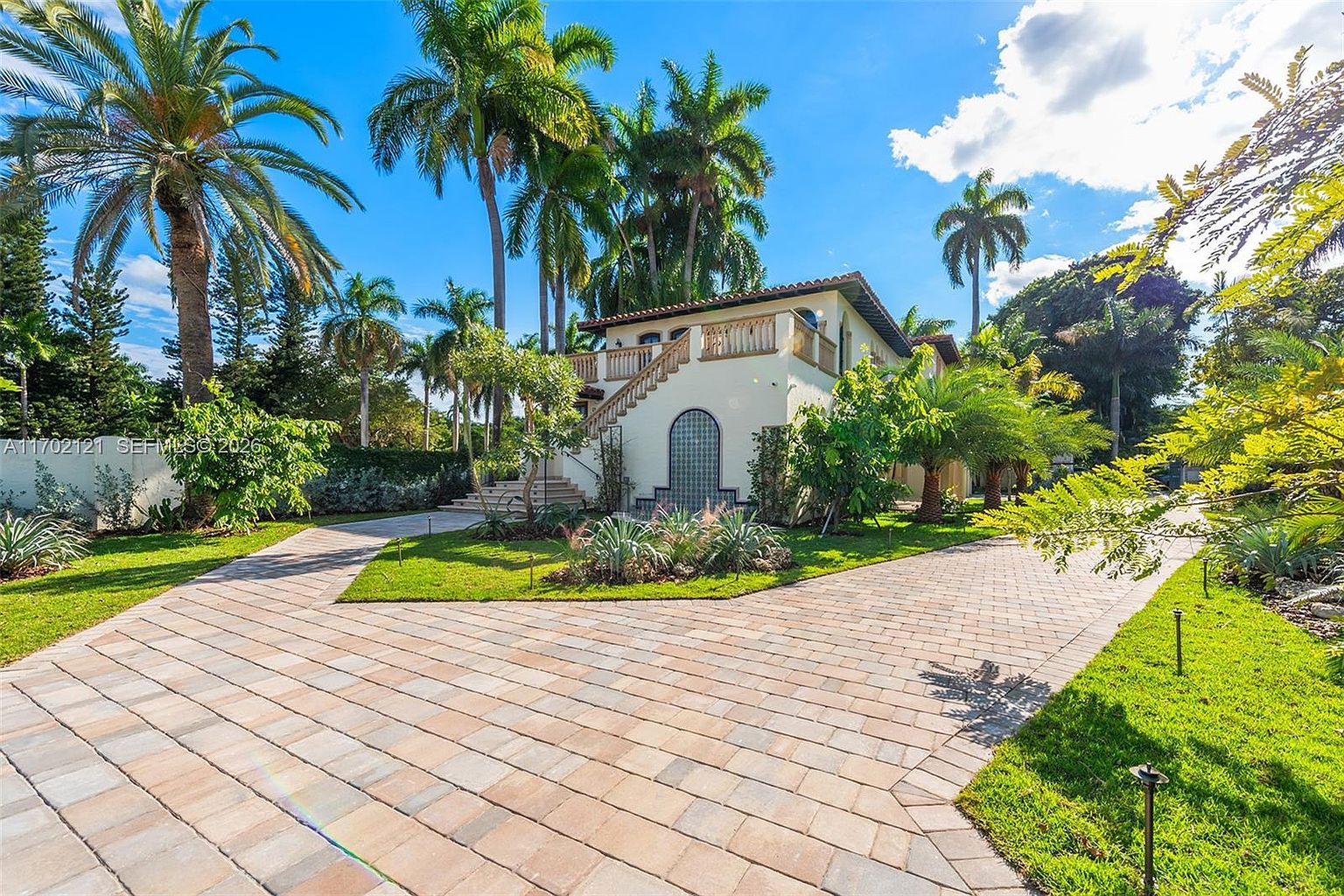 This Mediterranean-style home features a prominent exterior staircase leading to an upper-level balcony, set against a backdrop of lush tropical landscaping and tall palm trees. A wide, paved driveway leads toward the house, which is accented by an arched entryway and decorative tile work. The bright, sunny atmosphere and well-manicured lawn create an inviting and upscale curb appeal.
