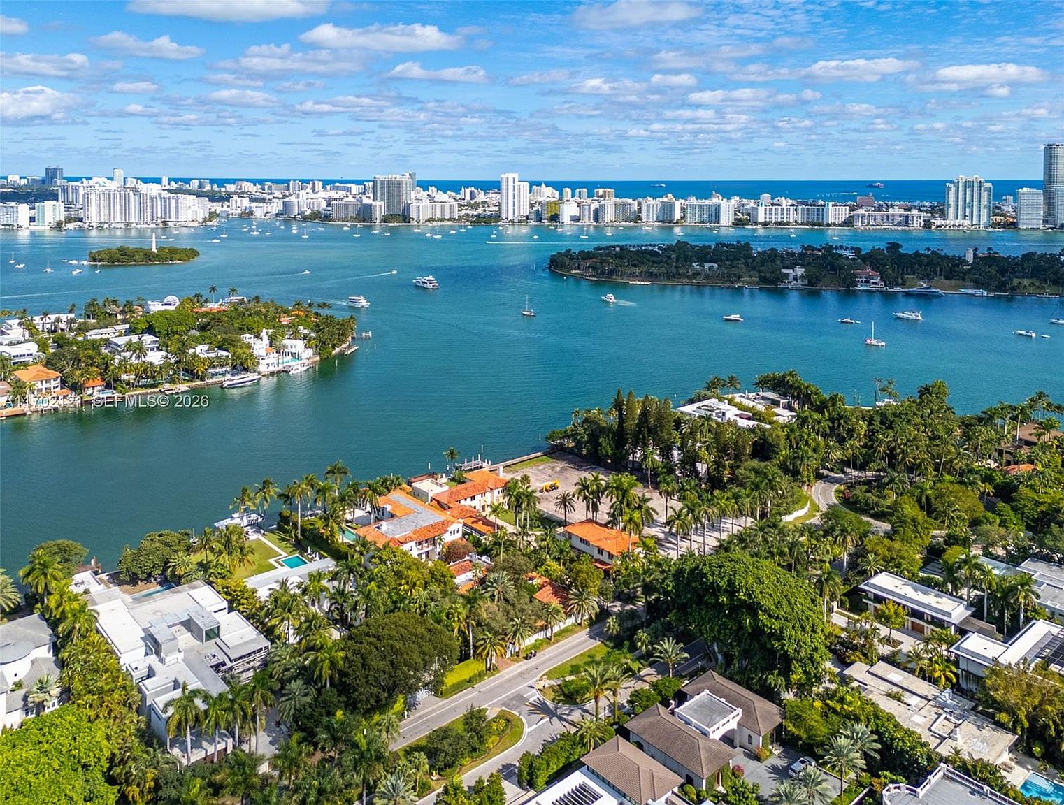 This high-angle aerial view captures a luxurious waterfront property nestled among lush tropical landscaping and mature trees. The perspective showcases the expansive bay, dotted with boats and islands, leading to a distant city skyline under a bright, partly cloudy sky. The scene emphasizes the prime coastal location, highlighting the integration of residential estates with the surrounding natural waterways.