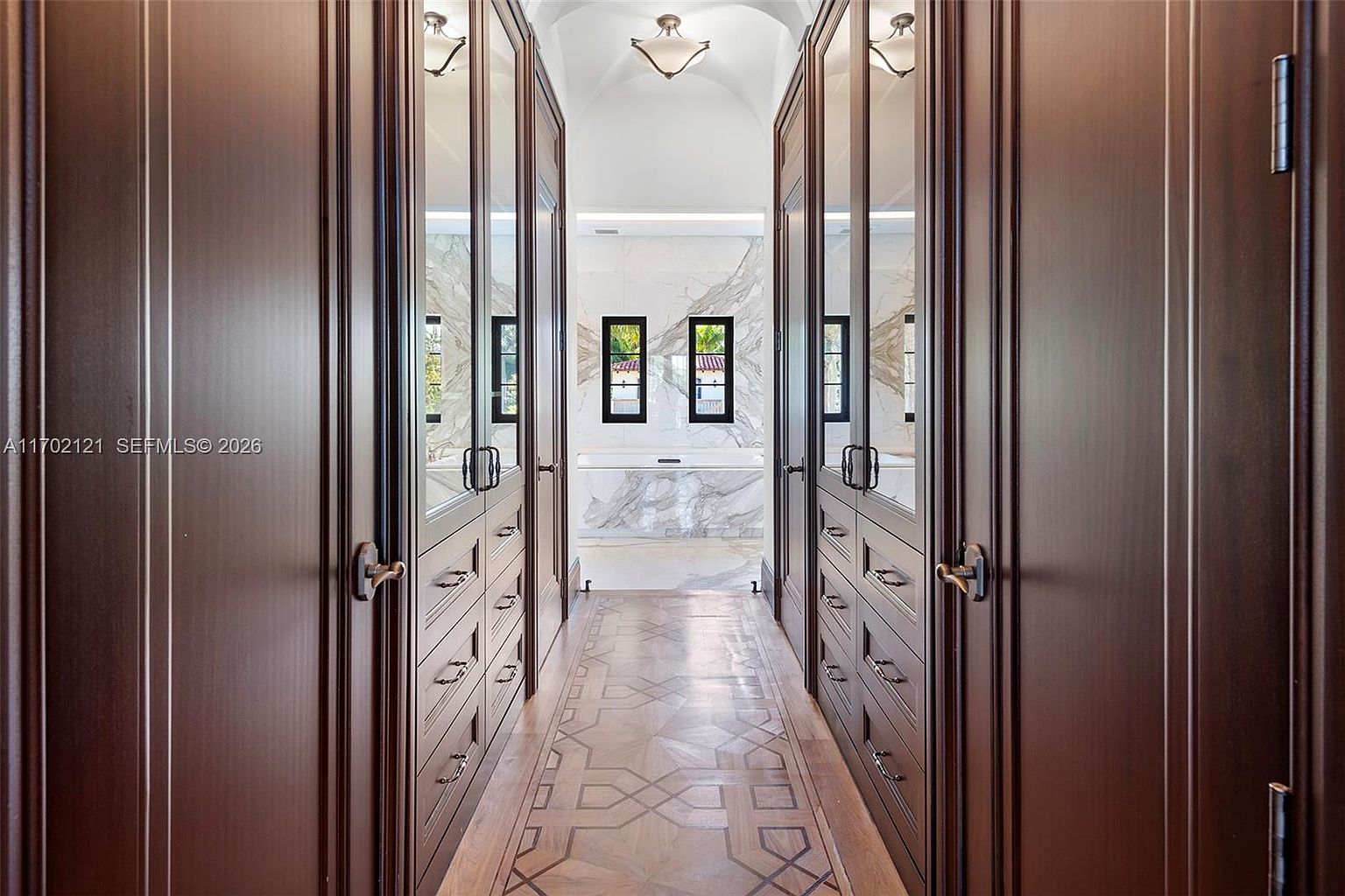 This elegant walk-in closet features dark wood cabinetry with mirrored door panels lining both sides of a central corridor. The space leads toward a bright bathroom area with a stone-clad wall and a soaking tub visible through the opening. The floor is adorned with intricate geometric wood inlay, creating a sophisticated and luxurious atmosphere.