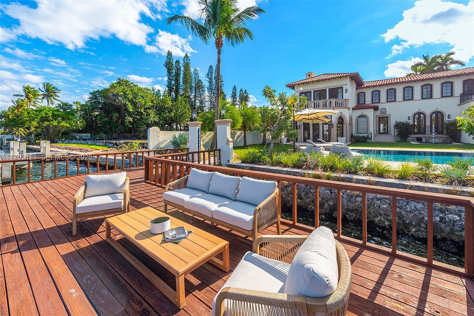 This expansive wooden deck overlooks a serene waterfront, featuring a comfortable outdoor seating area with a sofa, armchairs, and a central coffee table. Beyond the deck, a lush green lawn leads to a swimming pool and a large, Mediterranean-style residence with a tiled roof. The scene is captured from a slightly elevated perspective, emphasizing the seamless transition between the private deck and the resort-like backyard.