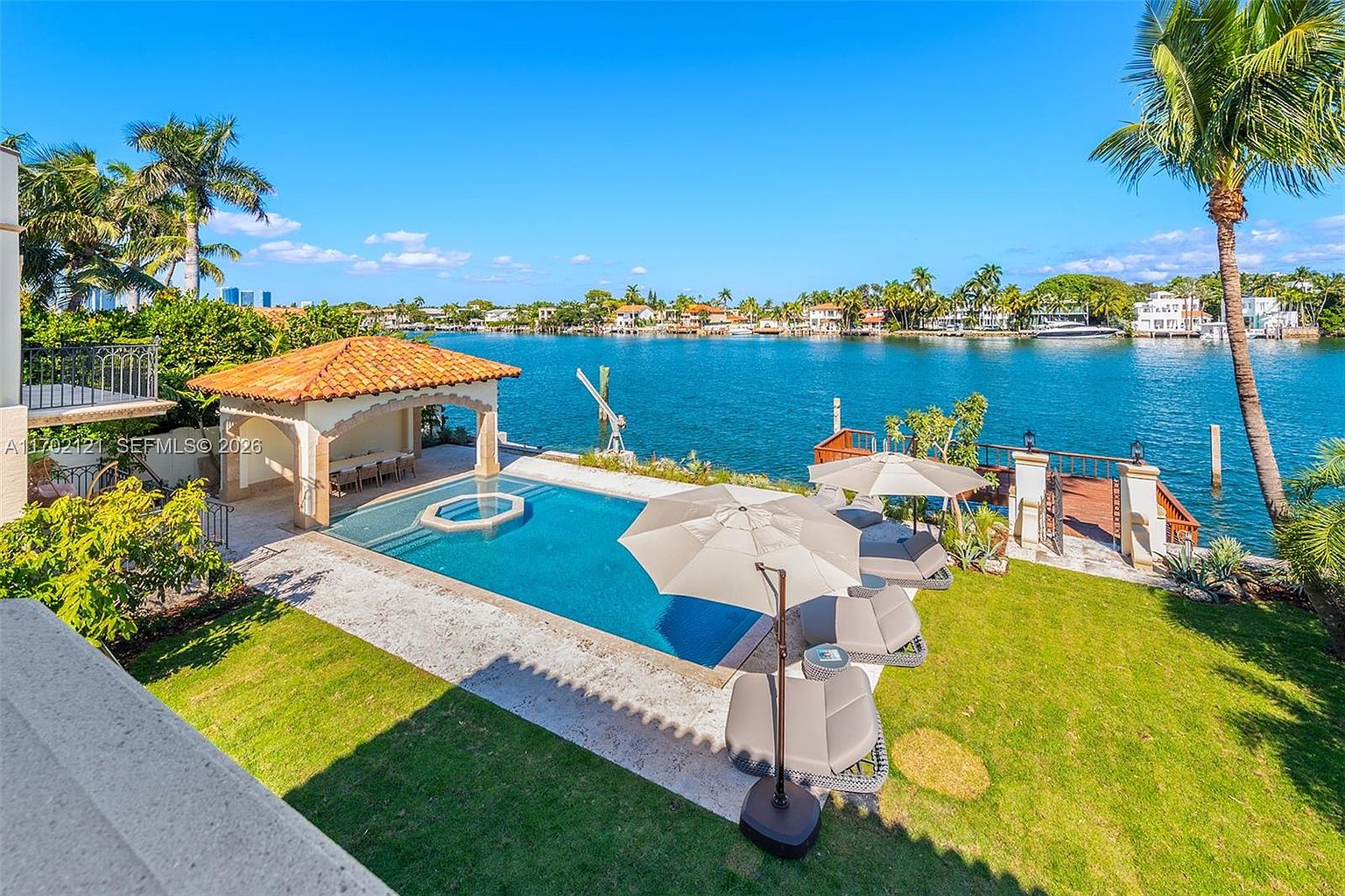 This elevated perspective captures a luxurious waterfront backyard featuring a rectangular swimming pool with an integrated spa. A covered pavilion with dining space sits adjacent to the pool, while a grassy lawn area is furnished with lounge chairs and umbrellas. The property overlooks a calm waterway, creating a serene and high-end outdoor living environment.
