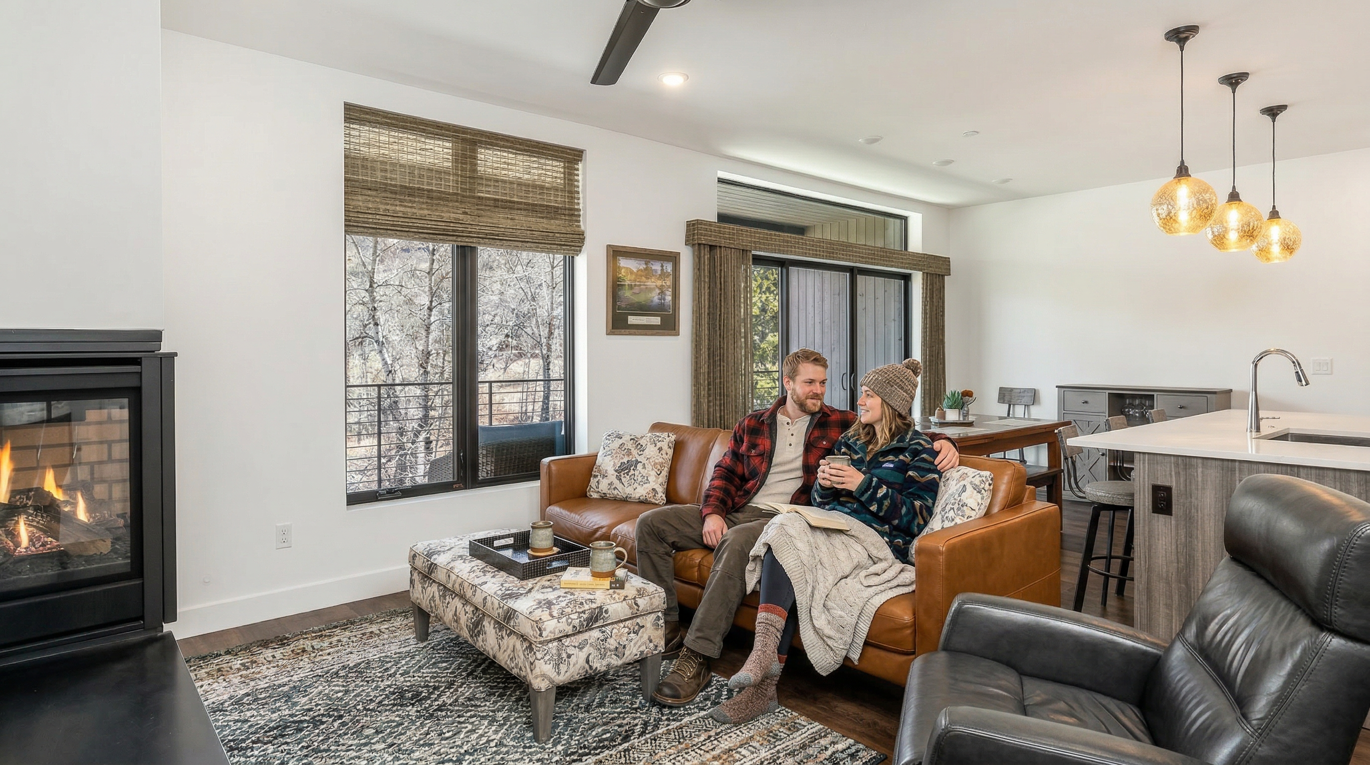 This inviting, open-concept living area features a cozy brown leather sofa and a patterned ottoman, perfectly centered around a modern fireplace. The space is anchored by a textured rug and finished with charming accents like woven blinds, warm pendant lighting, and a neutral, airy design palette. It provides a comfortable and stylish setting, ideal for relaxed living and casual entertaining.