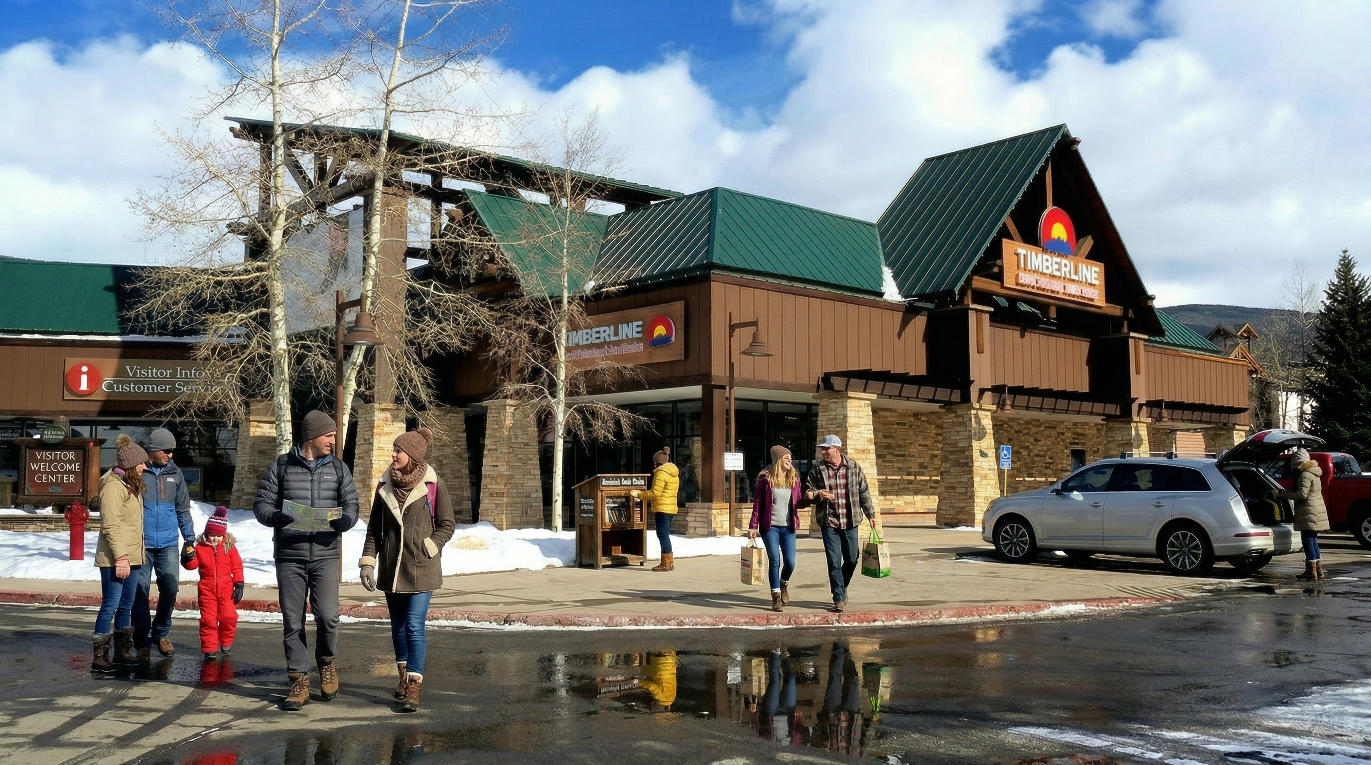 This image shows a commercial shopping center, specifically the 'Timberline' plaza, featuring rustic wood and stone architecture consistent with a mountain resort community. The building has a prominent gabled roof with green metal shingles and provides visitor information services, creating an inviting, active atmosphere for guests and local shoppers.