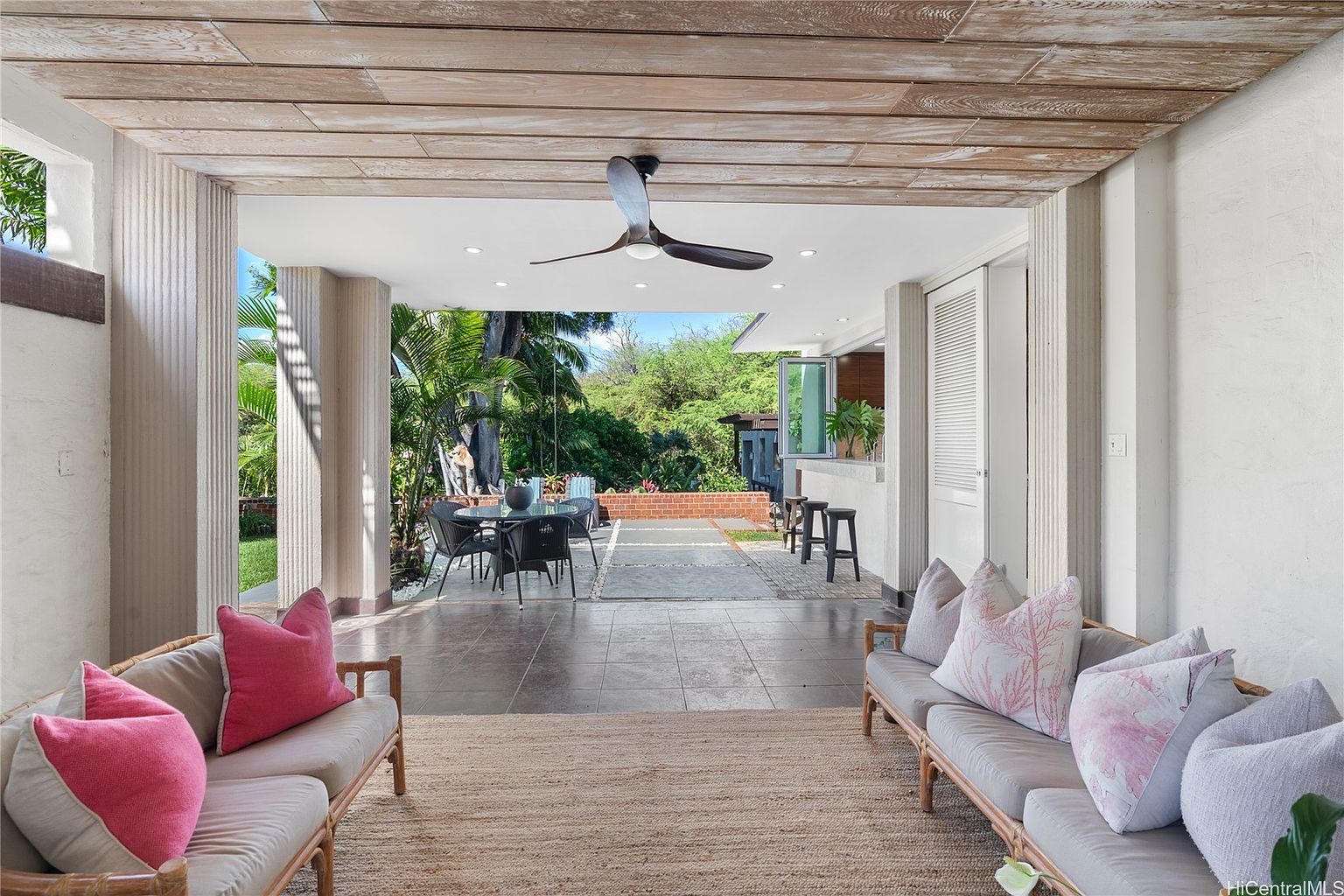 This image showcases a covered patio area, seamlessly blending indoor and outdoor living. The space features comfortable seating arrangements with plush cushions, a ceiling fan for air circulation, and an outdoor dining set visible in the background. The design incorporates natural elements like wood and stone, creating a relaxed and inviting atmosphere.
