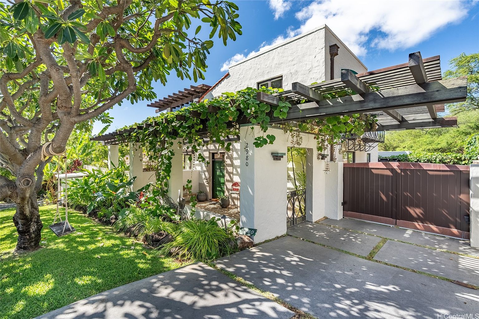 This is a charming front view of a two-story white stucco house with a lush, overgrown garden. A wooden pergola covered in vines extends from the house, creating a shaded entryway. A dark wooden gate provides privacy, and a mature tree with a swing adds character to the front yard.