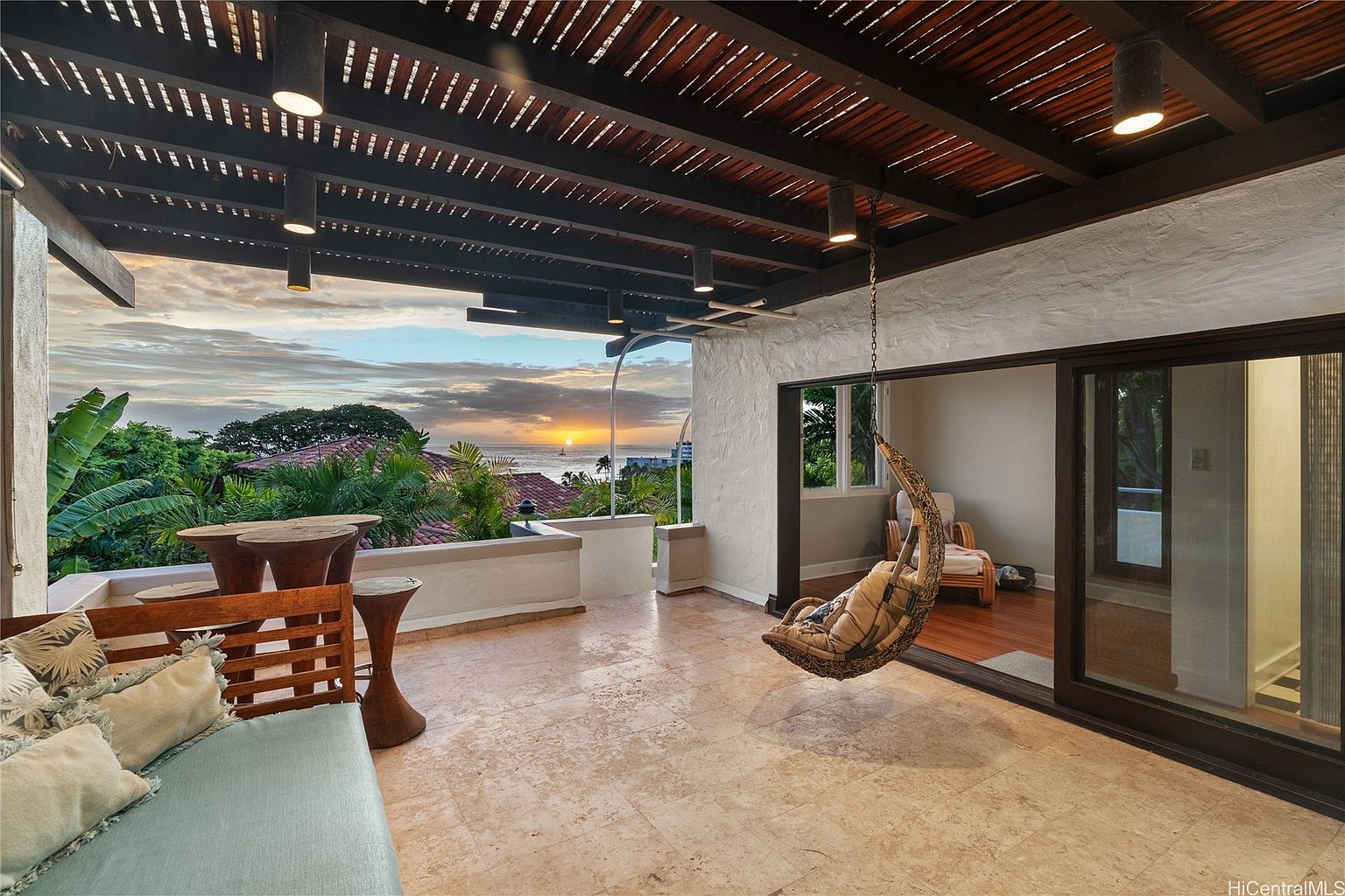 This image showcases a luxurious outdoor living space, featuring a spacious balcony with travertine flooring and a partial ocean view at sunset. The area is furnished with a wooden bench, unique side tables, and a hanging chair, creating a relaxing atmosphere. The transition to the interior is seamless with large sliding glass doors, highlighting the indoor-outdoor flow.