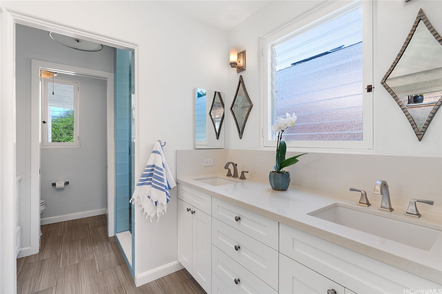 This is a bright and clean bathroom featuring a double vanity with white cabinetry and light countertops. Diamond-shaped mirrors and a modern light fixture add a touch of style. A shower with blue tiling is visible through an open doorway, and the flooring appears to be wood-look tile, creating a warm and inviting space.