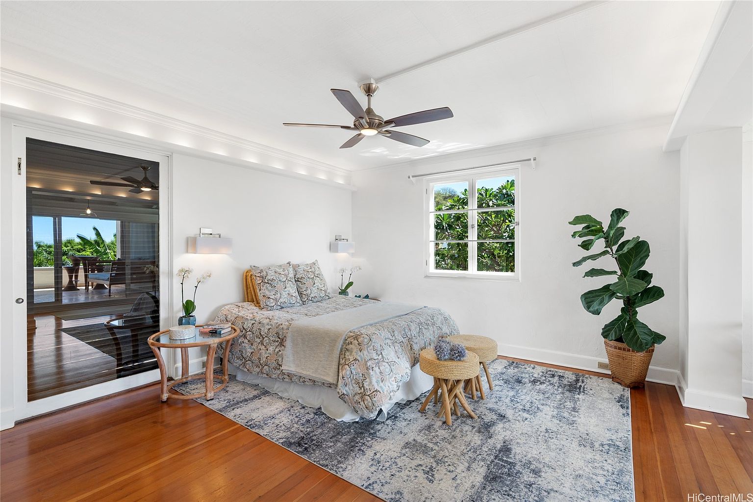 This is a bright and airy primary bedroom featuring hardwood floors, white walls, and a decorative ceiling. A large window provides natural light, complemented by wall-mounted sconces. The room includes a bed with patterned bedding, a small side table, and a potted plant, creating a serene and inviting atmosphere.