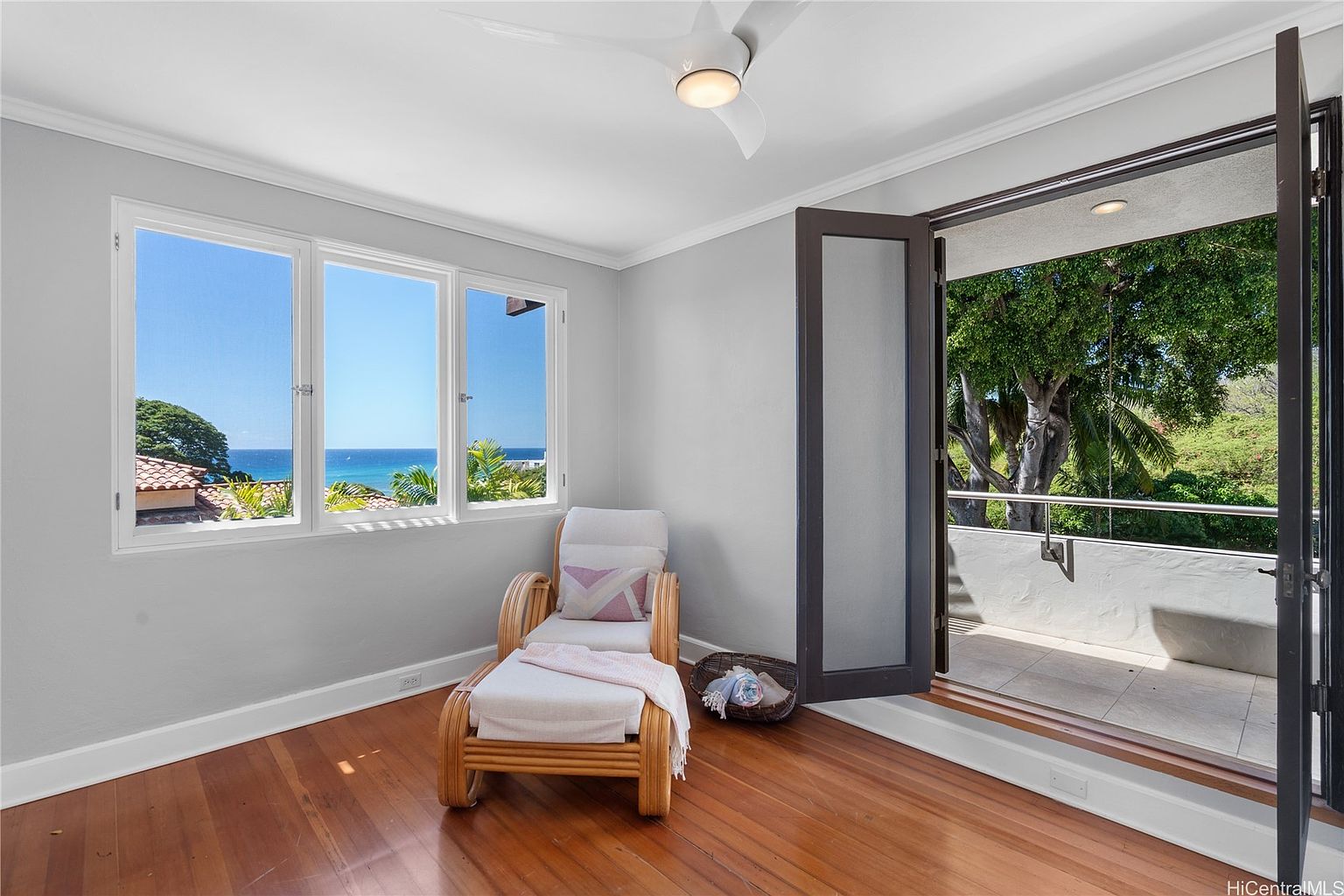 This is an interior shot of a bedroom featuring a relaxed seating area with a rattan chaise lounge, complemented by a view of the ocean through a set of windows. The room opens onto a balcony through dark-framed doors, inviting natural light and fresh air into the space, while the hardwood floors add warmth to the room.
