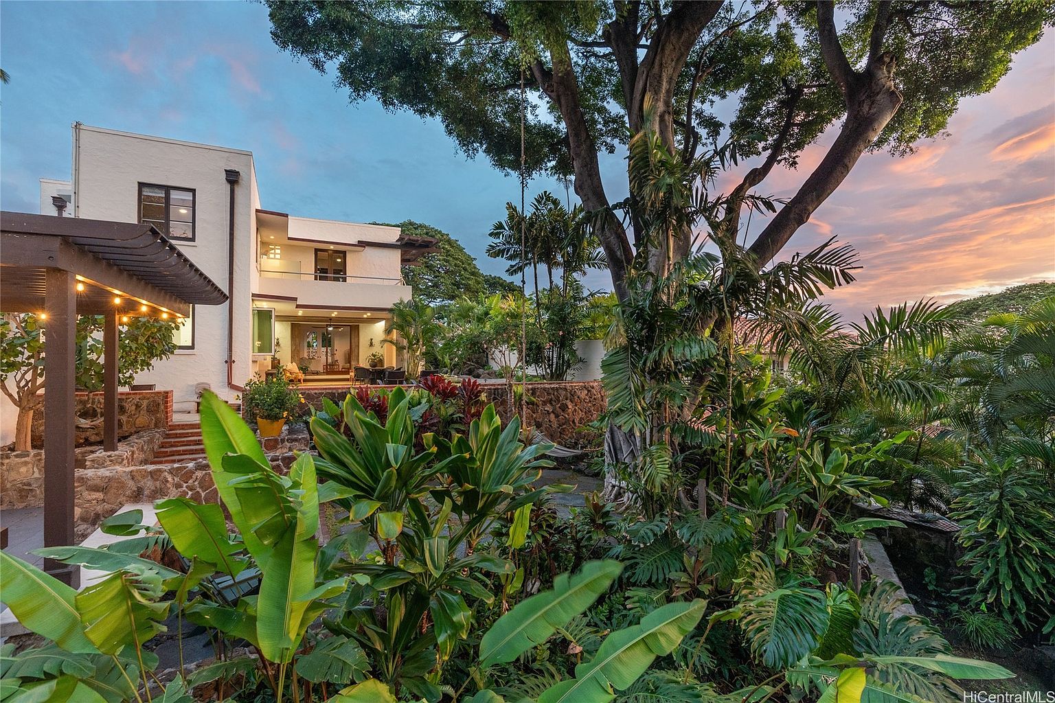 This image showcases a lush and vibrant yard/garden area, featuring a variety of tropical plants and trees. A multi-story house with a balcony is visible in the background, complemented by a stone patio area. The scene exudes a sense of tranquility and privacy, highlighting the property's outdoor living space.