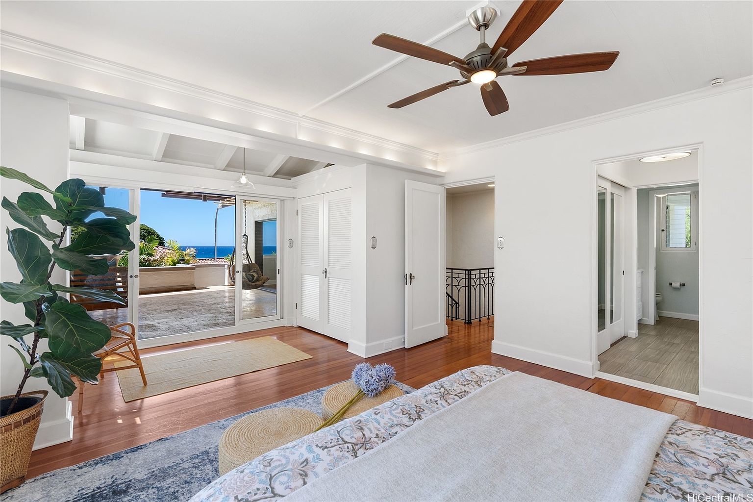 This is a bright and airy primary bedroom featuring hardwood floors, white walls, and a ceiling fan. A sliding glass door leads to a balcony with ocean views, enhancing the room's appeal. The room includes built-in closets and access to an ensuite bathroom, creating a luxurious and comfortable space.