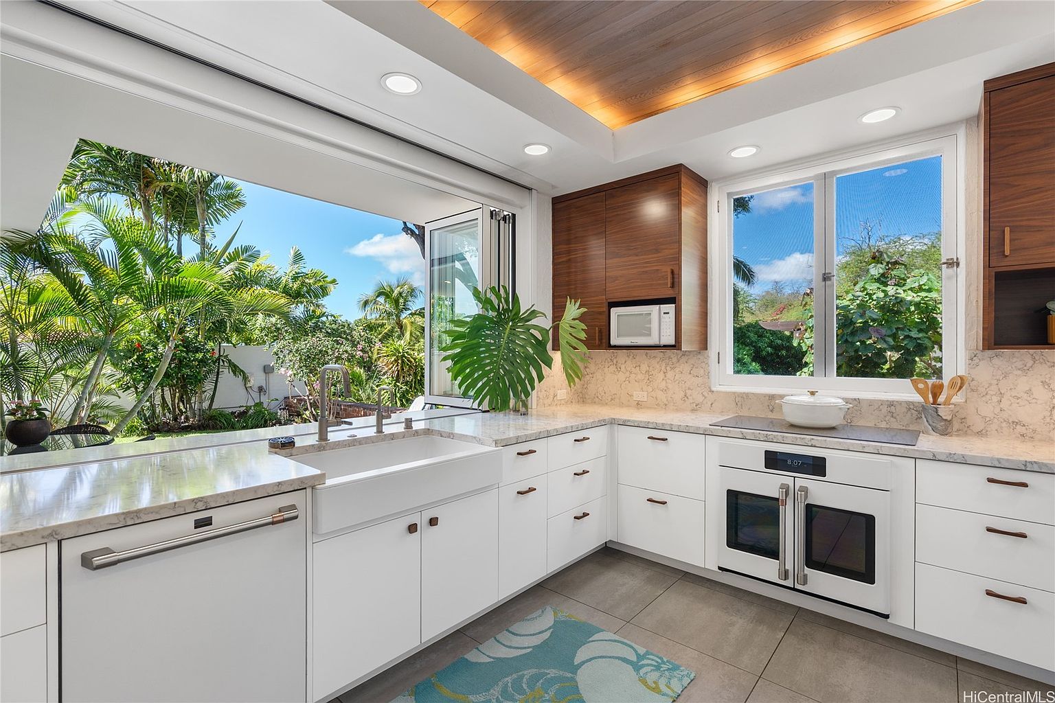 This is a bright and modern kitchen featuring white cabinetry, marble countertops, and stainless steel appliances. A large window above the sink opens to a lush tropical garden, bringing the outdoors in. The kitchen also features wood-paneled upper cabinets and recessed lighting, creating a warm and inviting atmosphere.