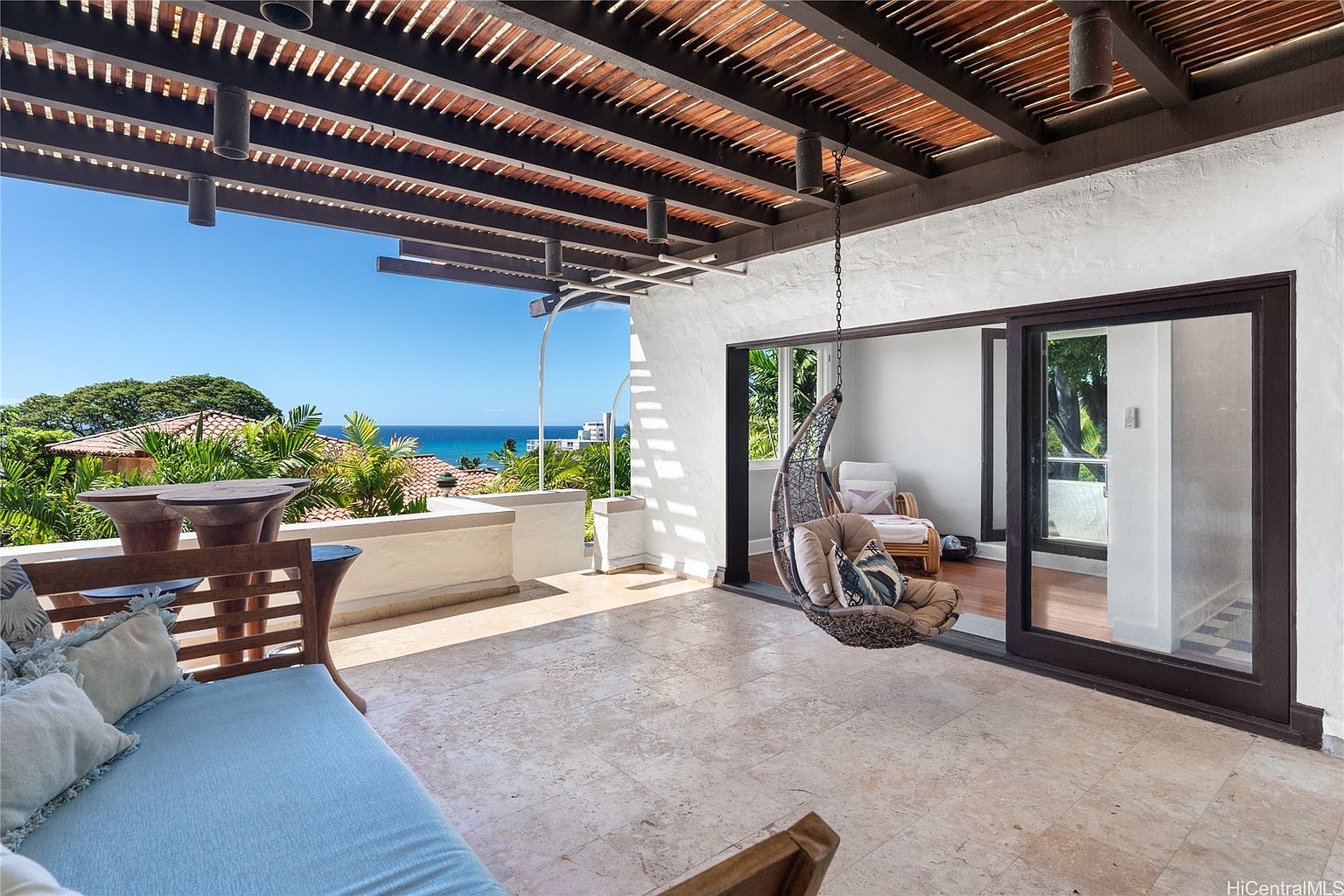 This image showcases a luxurious outdoor patio or balcony area with a stunning ocean view. The space features a comfortable seating area with a blue cushioned bench and a hanging chair, complemented by a stylish wooden ceiling structure. The flooring is tiled, and the area seamlessly connects to an interior space through sliding glass doors, creating an inviting indoor-outdoor living experience.