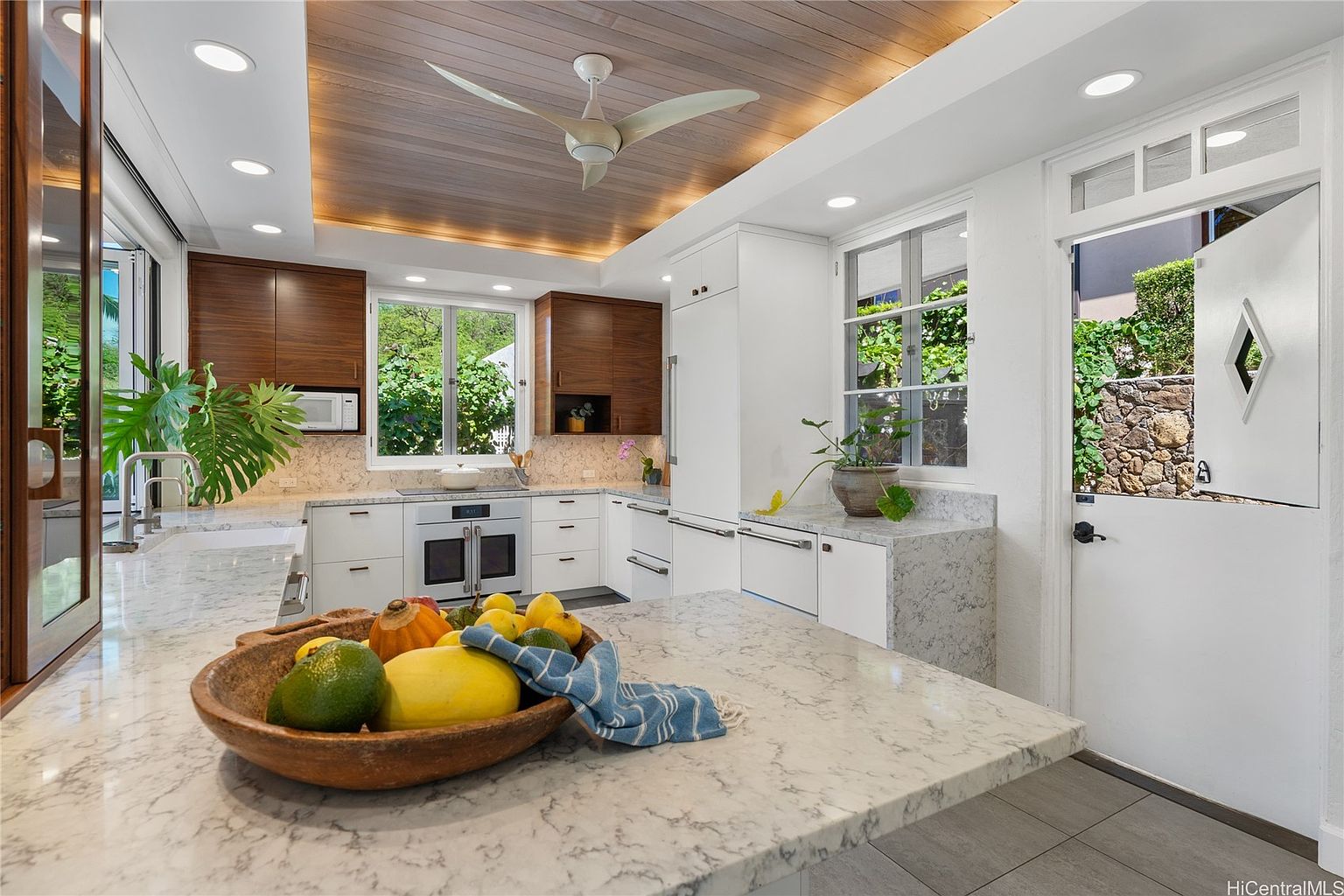 This is a bright and modern kitchen featuring a mix of white and wood cabinetry, complemented by light marble countertops. A large island takes center stage, adorned with a wooden bowl of fruit, while stainless steel appliances add a touch of elegance. Natural light floods the space through large windows, creating a warm and inviting atmosphere.