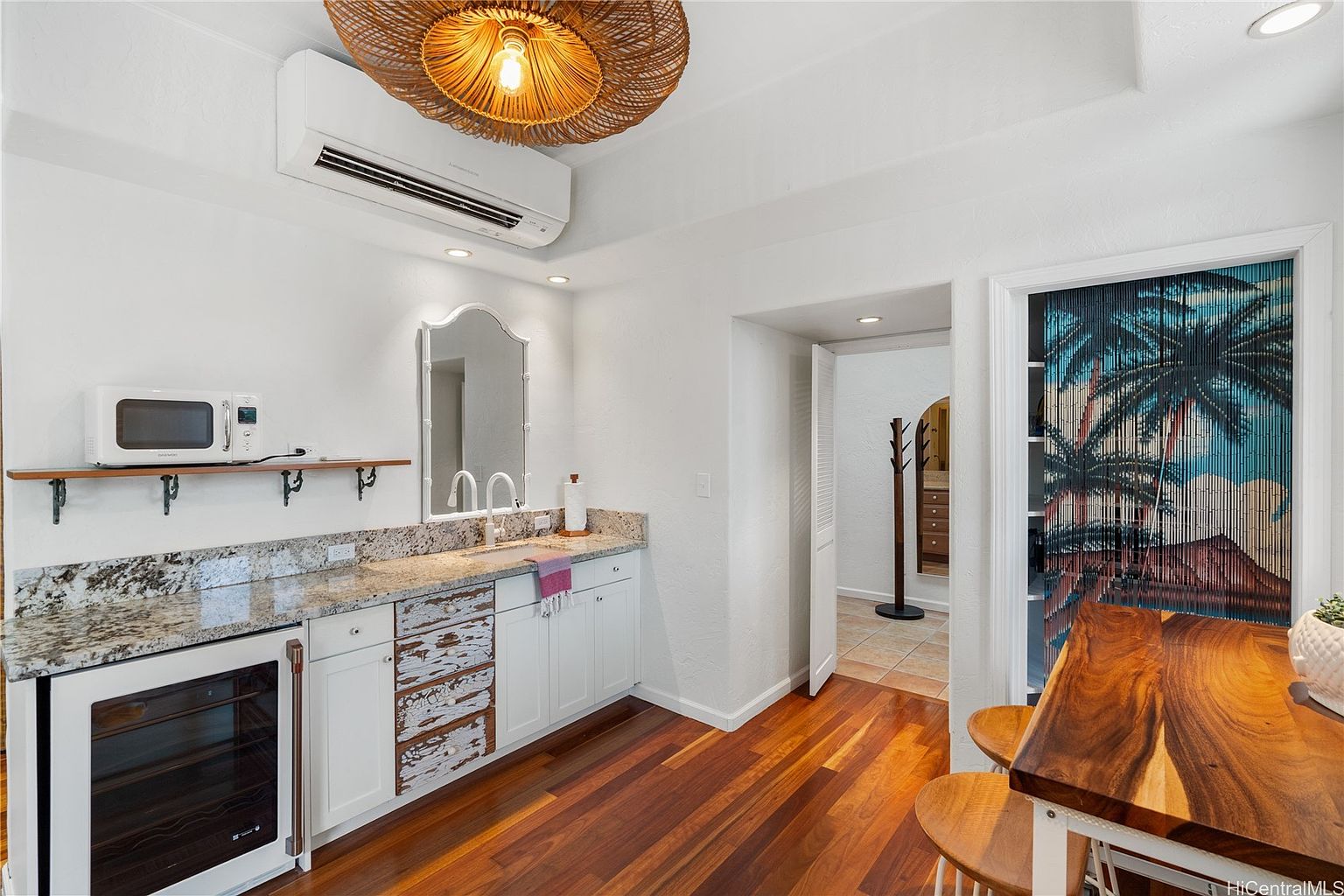 This interior shot showcases a well-lit kitchen area with hardwood floors and white cabinetry. A granite countertop complements the cabinets, and a microwave sits on a shelf above. The room features a unique light fixture and a doorway leading to another room, adding depth and character to the space.