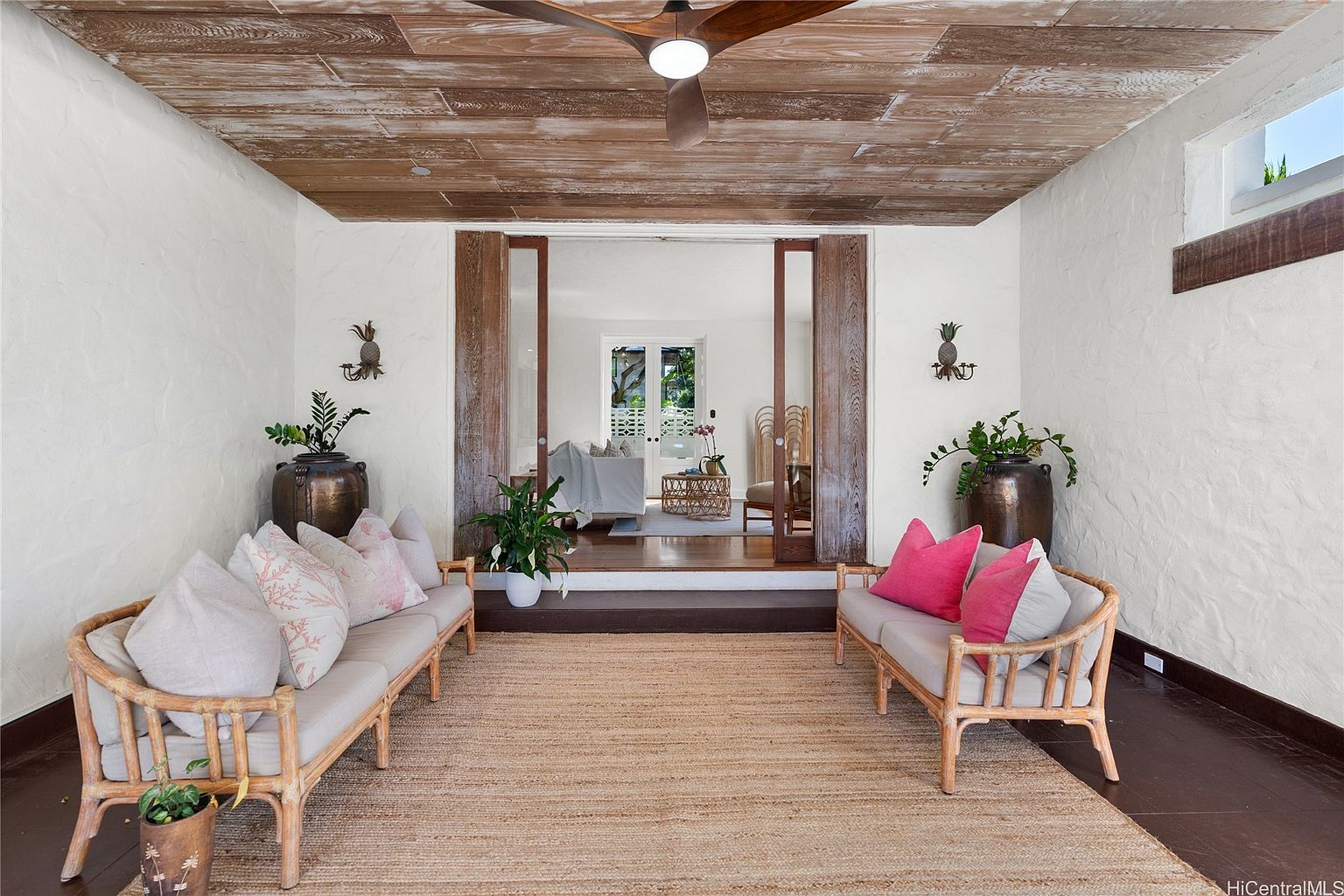 This sunroom features a rustic-chic design with wooden plank ceilings and textured white walls. Two rattan sofas with decorative pillows flank a jute rug, creating a cozy seating area. The open doorway leads to another room, enhancing the sense of spaciousness and connection between spaces.