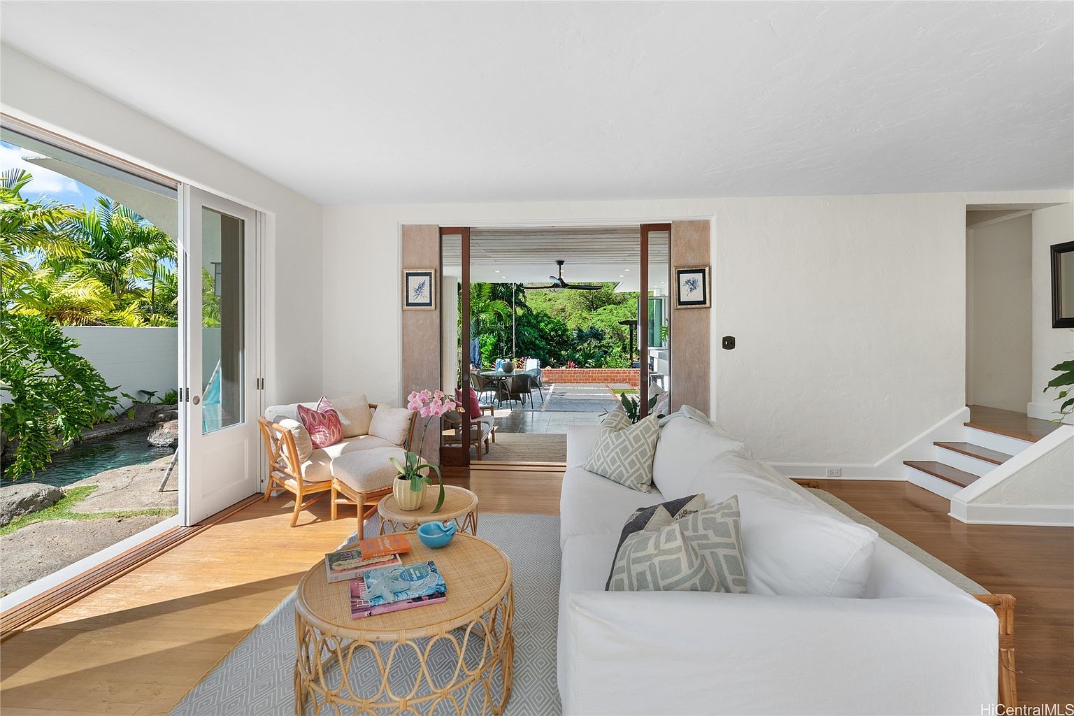 This is an interior shot of a bright and airy living room. The room features a white sofa with patterned pillows, a rattan coffee table, and a cozy seating area with a view of a small pond through a sliding glass door. The open floor plan leads to an outdoor patio, creating a seamless indoor-outdoor living experience.
