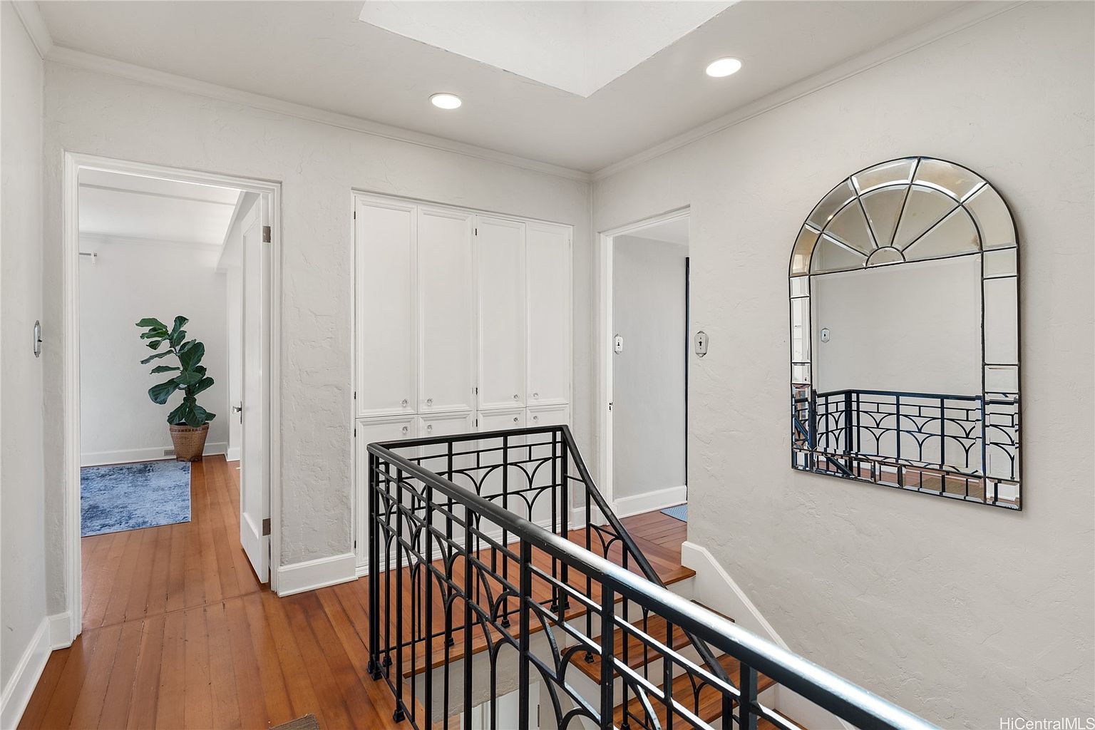 This interior shot showcases a well-lit hallway and staircase. The hallway features hardwood floors, white walls, and built-in storage cabinets. A decorative mirror hangs on the wall, reflecting the staircase with its black wrought iron railing, creating an elegant and inviting transition space within the home.