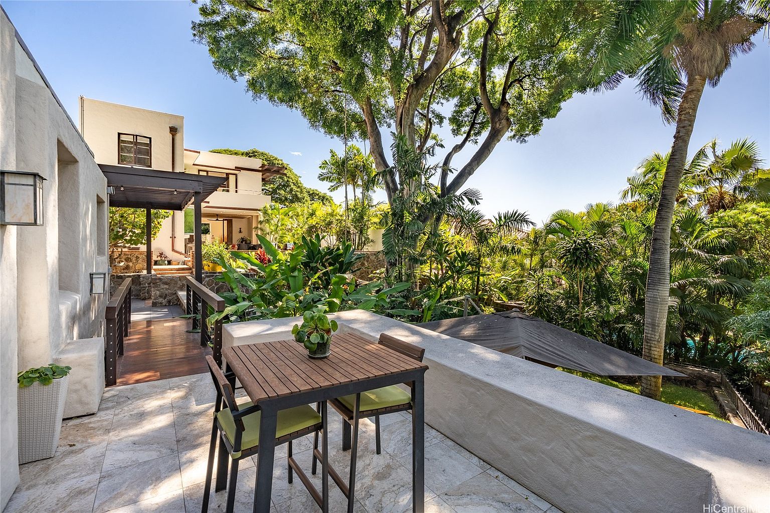 This image showcases a charming outdoor patio area, featuring a wooden table and chairs set on a tiled surface. Lush greenery and mature trees surround the space, creating a private and serene atmosphere. The patio is connected to the main house via a wooden walkway, adding to the property's appeal.