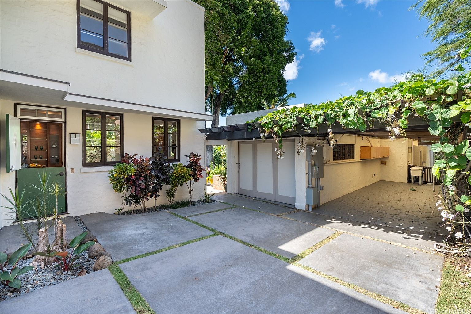 This exterior side view showcases a charming property with a concrete driveway featuring grass inlays, leading to a covered carport adorned with lush vines. The building's facade is painted in a bright white, complemented by dark-framed windows and a unique green Dutch door, adding character and curb appeal. The landscaping includes a mix of colorful shrubs and small trees, enhancing the overall aesthetic.