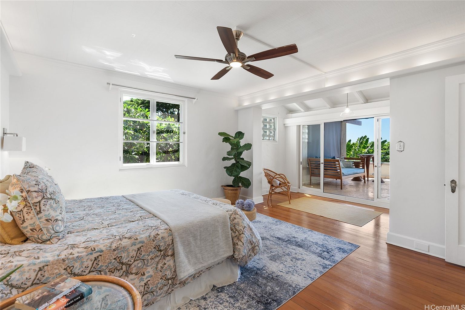 This is a bright and airy primary bedroom featuring hardwood floors, white walls, and a ceiling fan. A large window provides natural light and a view of greenery, while sliding glass doors lead to an outdoor patio or balcony. The room is furnished with a bed, a rug, and a wicker chair, creating a comfortable and inviting space.